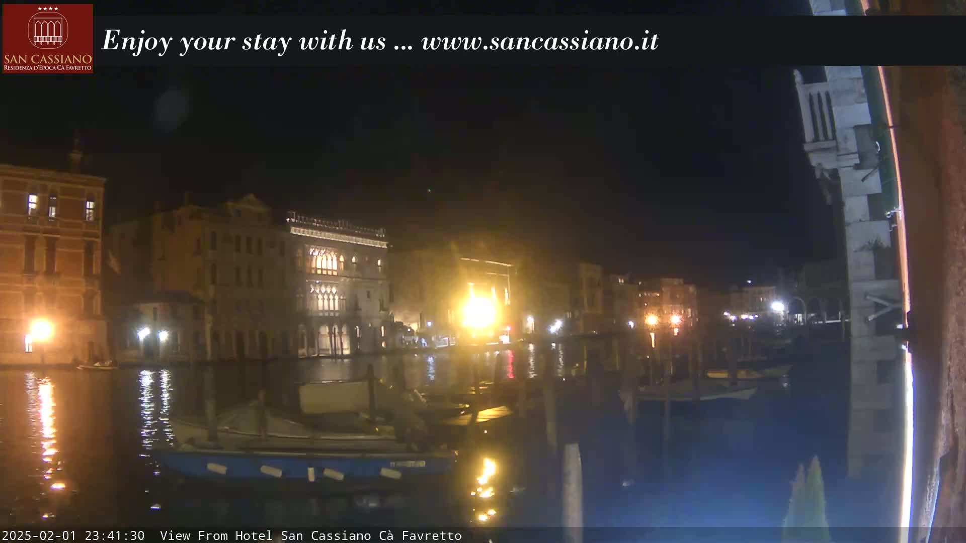 A nighttime view of a canal in Venice, with several buildings illuminated along its banks, shows boats moored on the water.