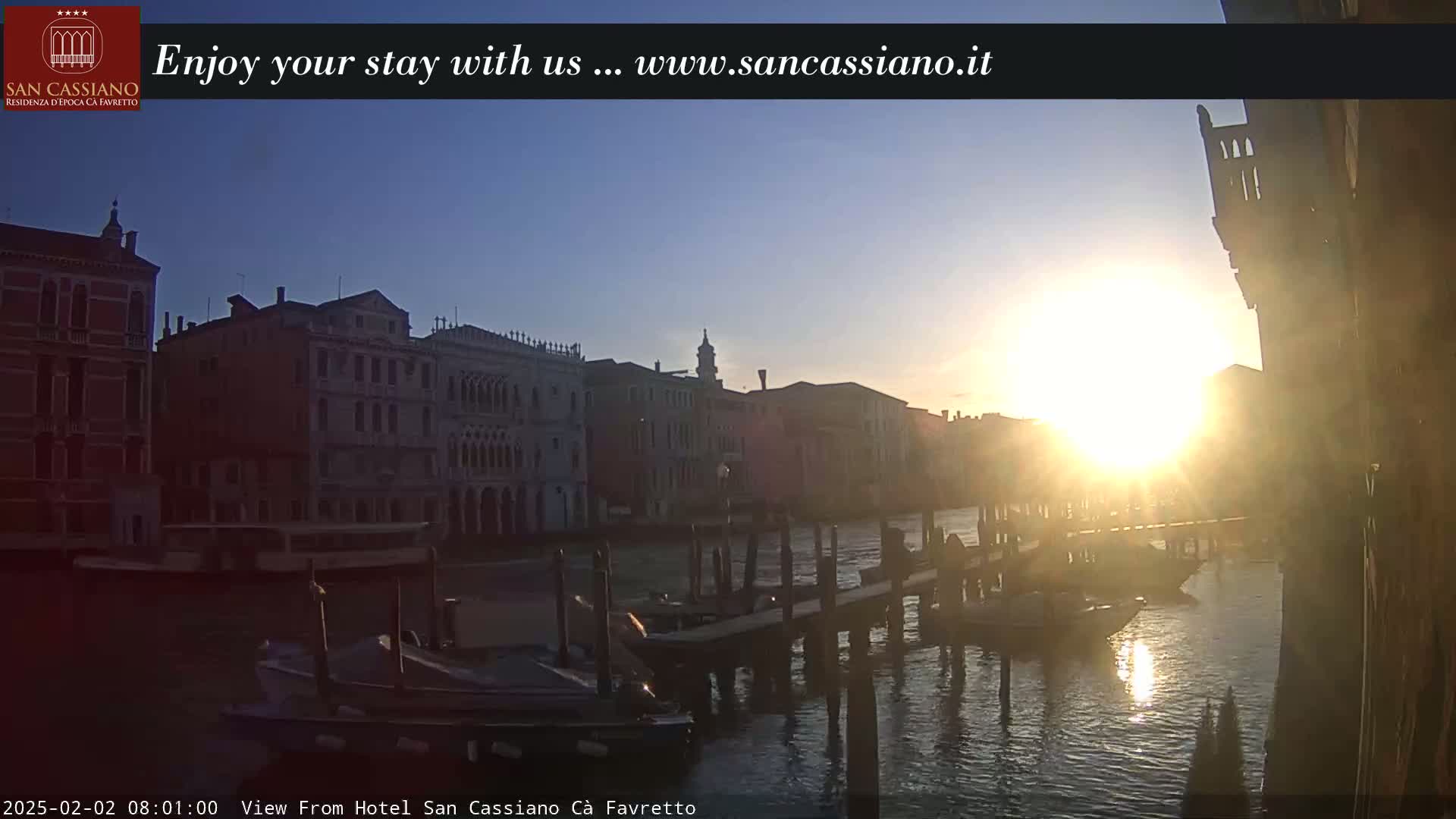 A sunrise illuminates a Venetian canal, reflecting off the water and silhouetting buildings and boats along the waterfront.