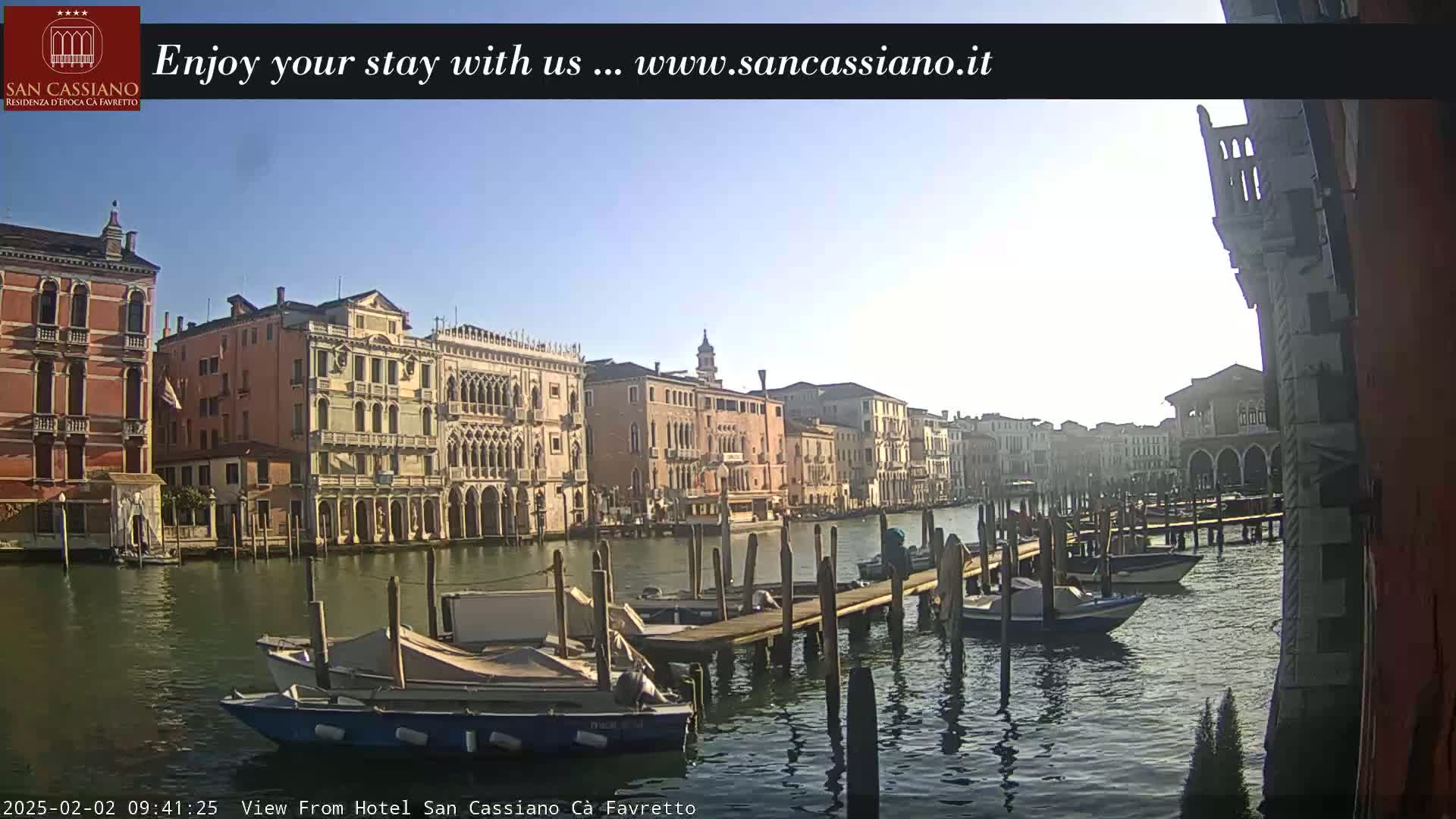 A sunny day in Venice shows several boats moored at a wooden dock along a canal lined with historic buildings.