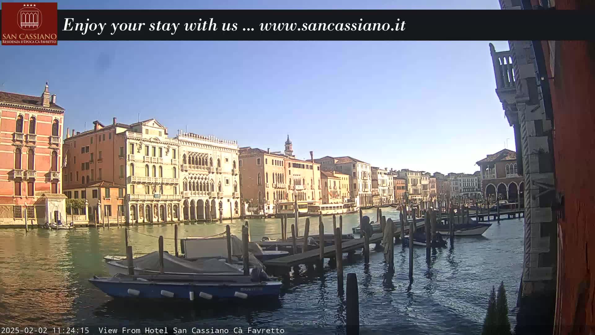 A sunny day in Venice shows a canal with several boats moored at a dock, and numerous historic buildings lining the waterway.