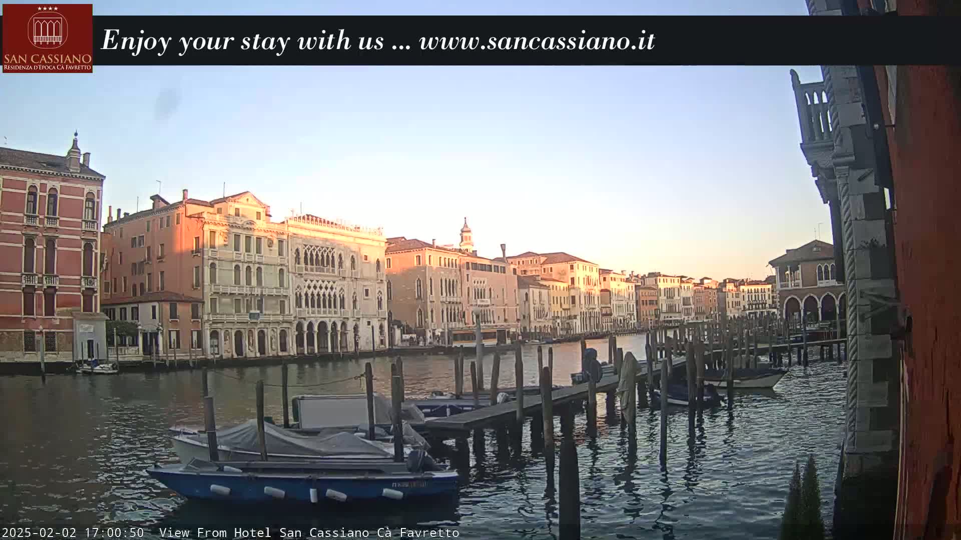 Several boats are moored at a dock along a canal lined with Venetian buildings under a clear, twilight sky.
