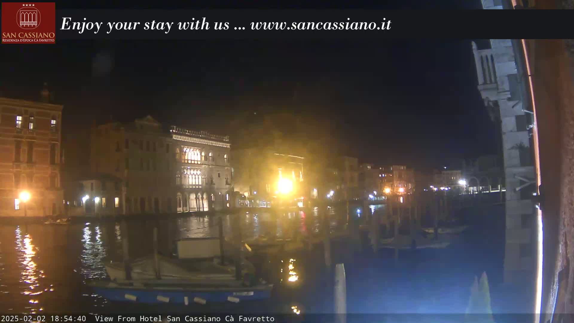 A nighttime view across a canal shows illuminated buildings along the waterfront, with boats moored nearby.