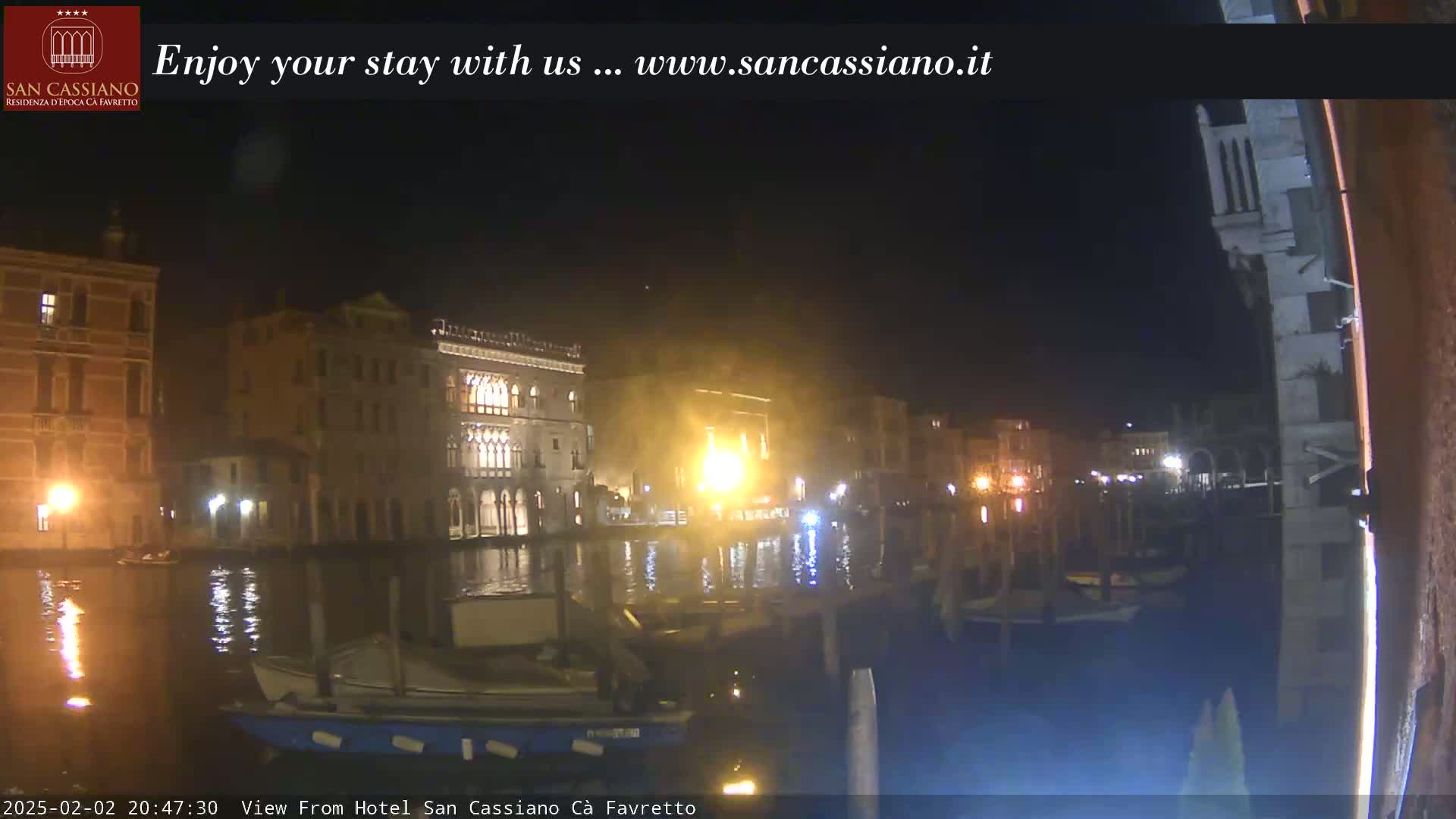 A nighttime view of a canal in Venice, showing several buildings illuminated along the water's edge with boats moored nearby.