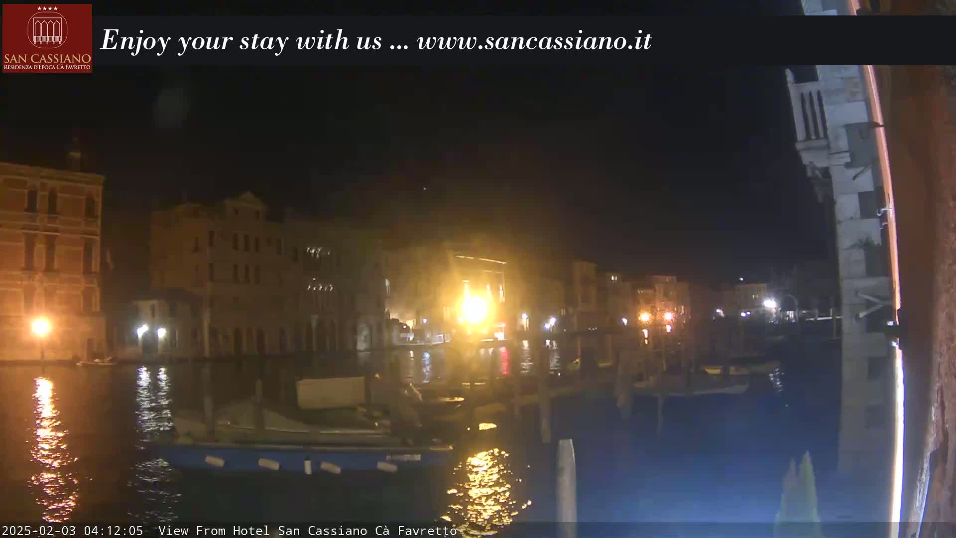A nighttime view of a canal in Venice, showing illuminated buildings and moored boats on calm water.