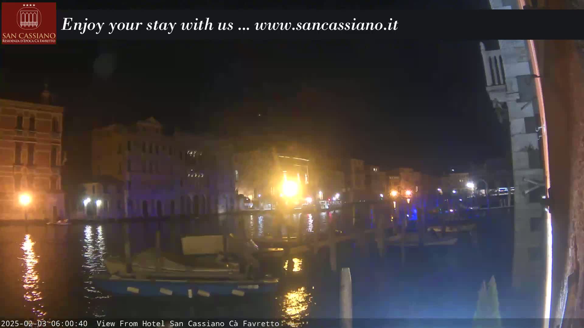 At night, a canal scene is shown with buildings lit up along its banks, boats on the water, and calm water reflecting the lights.