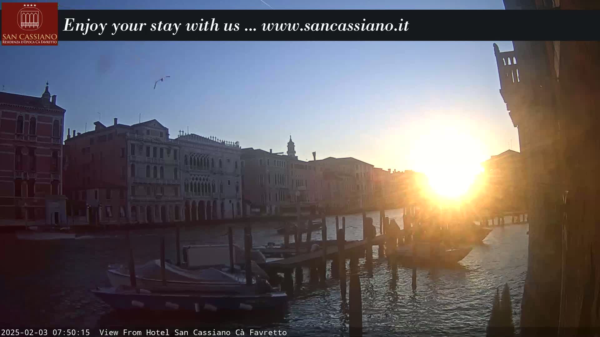 A sunrise illuminates a canal in Venice, with several boats moored along a wooden dock, and tall buildings lining the waterway.