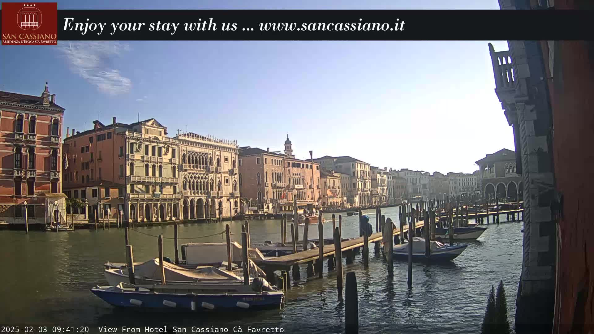 A sunny day in Venice shows several boats moored at a dock along a canal lined with historic buildings.