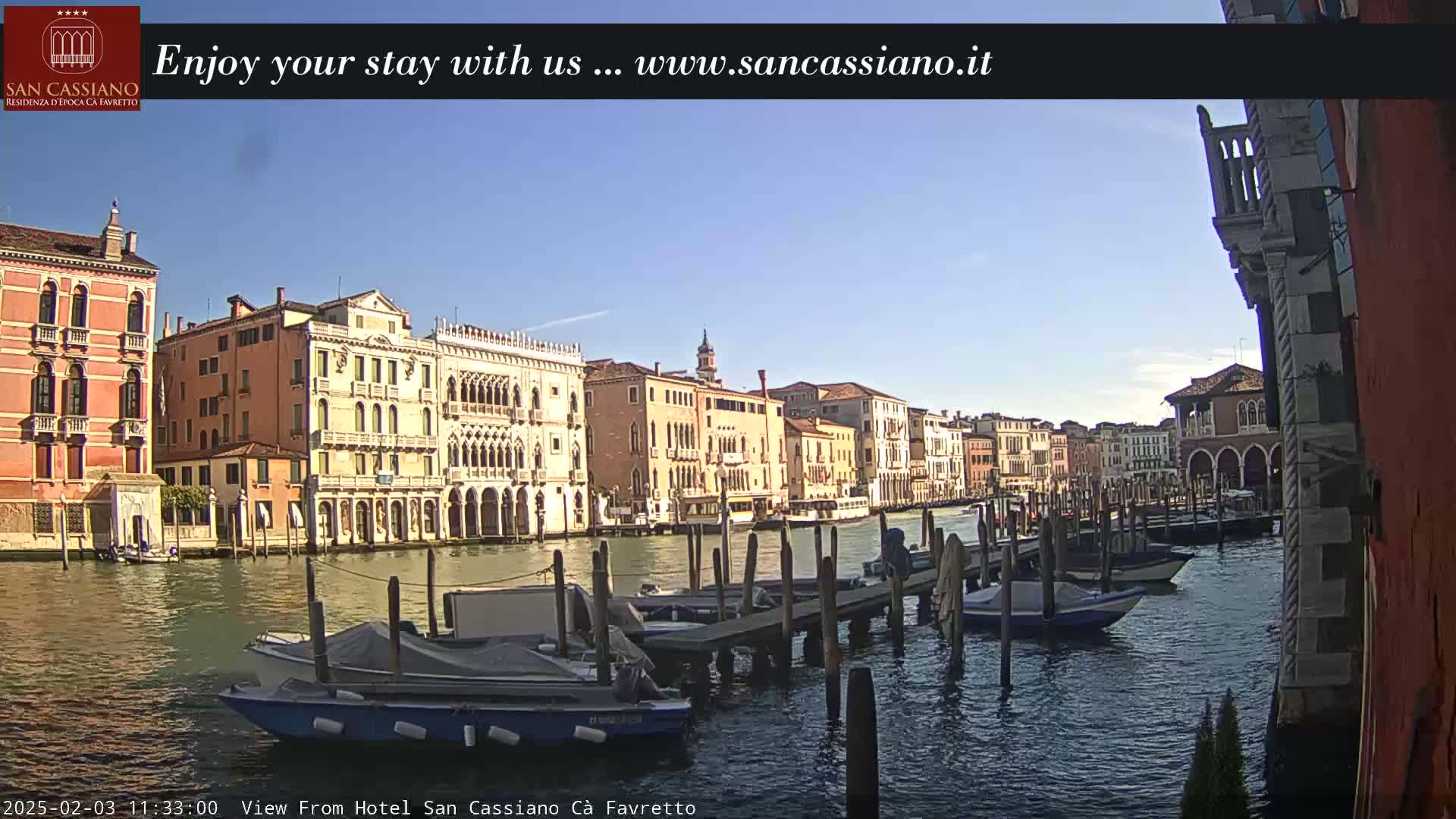 A sunny day in Venice shows colorful buildings lining a canal, with several boats moored at a dock.