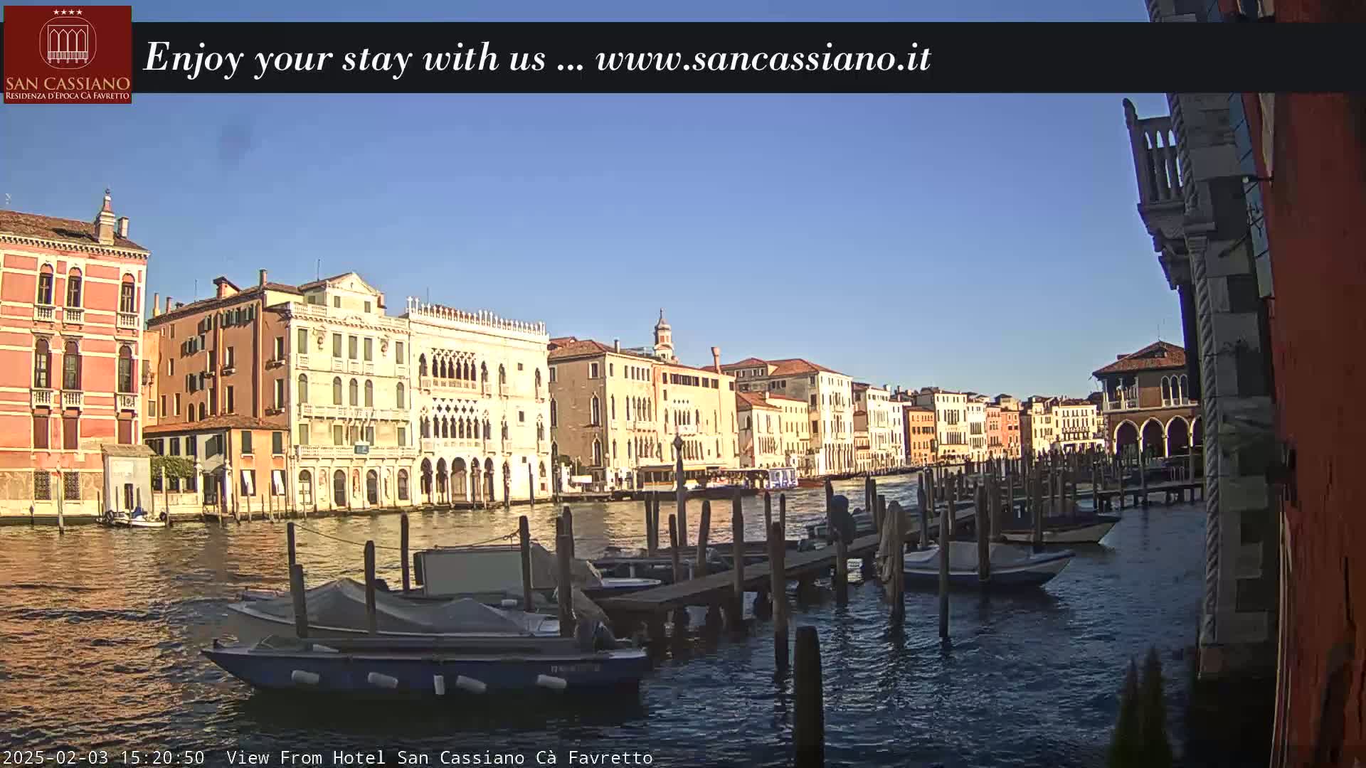 A sunny day in Venice shows a canal lined with historic buildings and several boats moored at a dock.