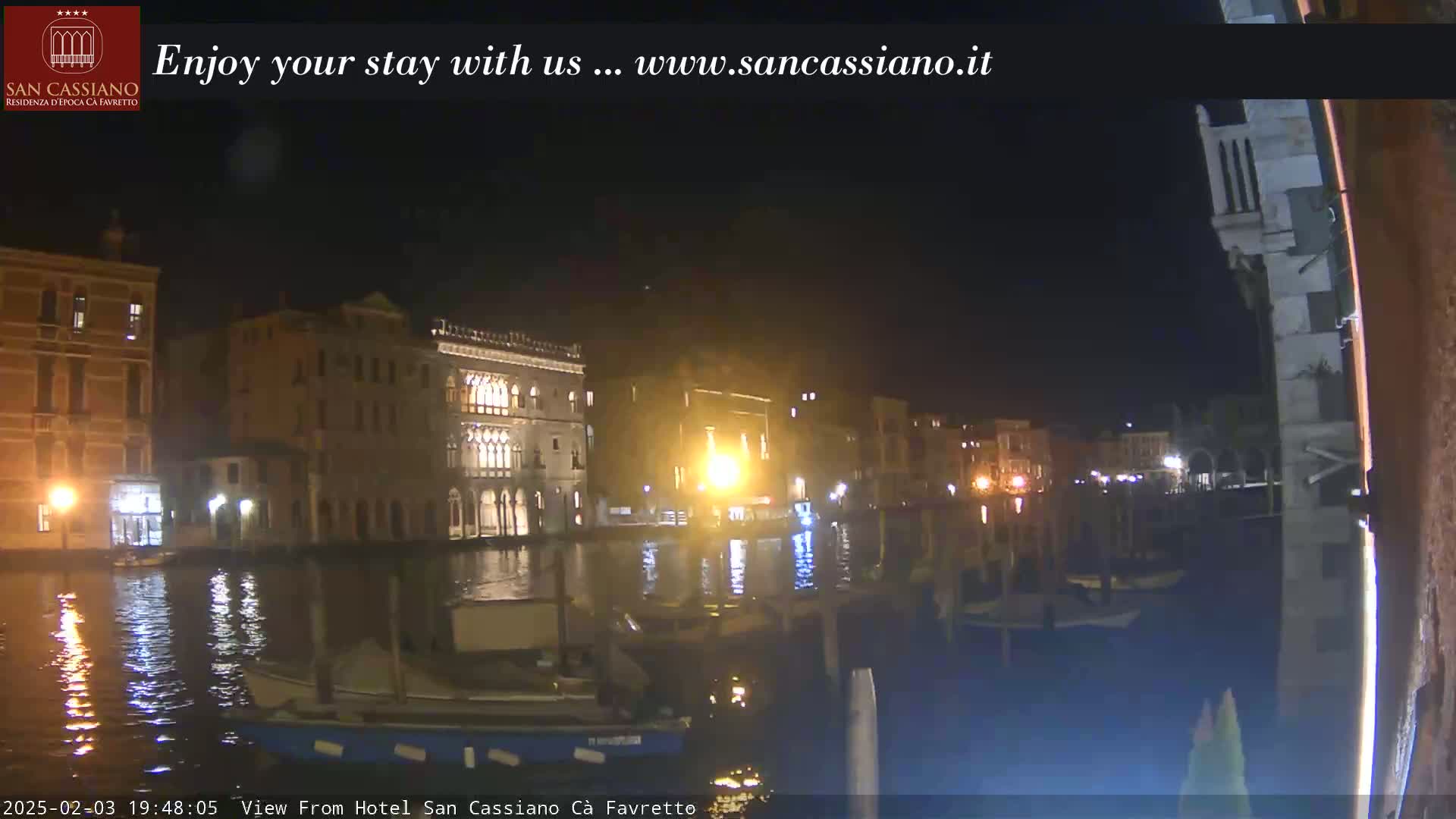 A nighttime view of a canal in Venice, showing illuminated buildings along the water's edge and boats moored nearby.
