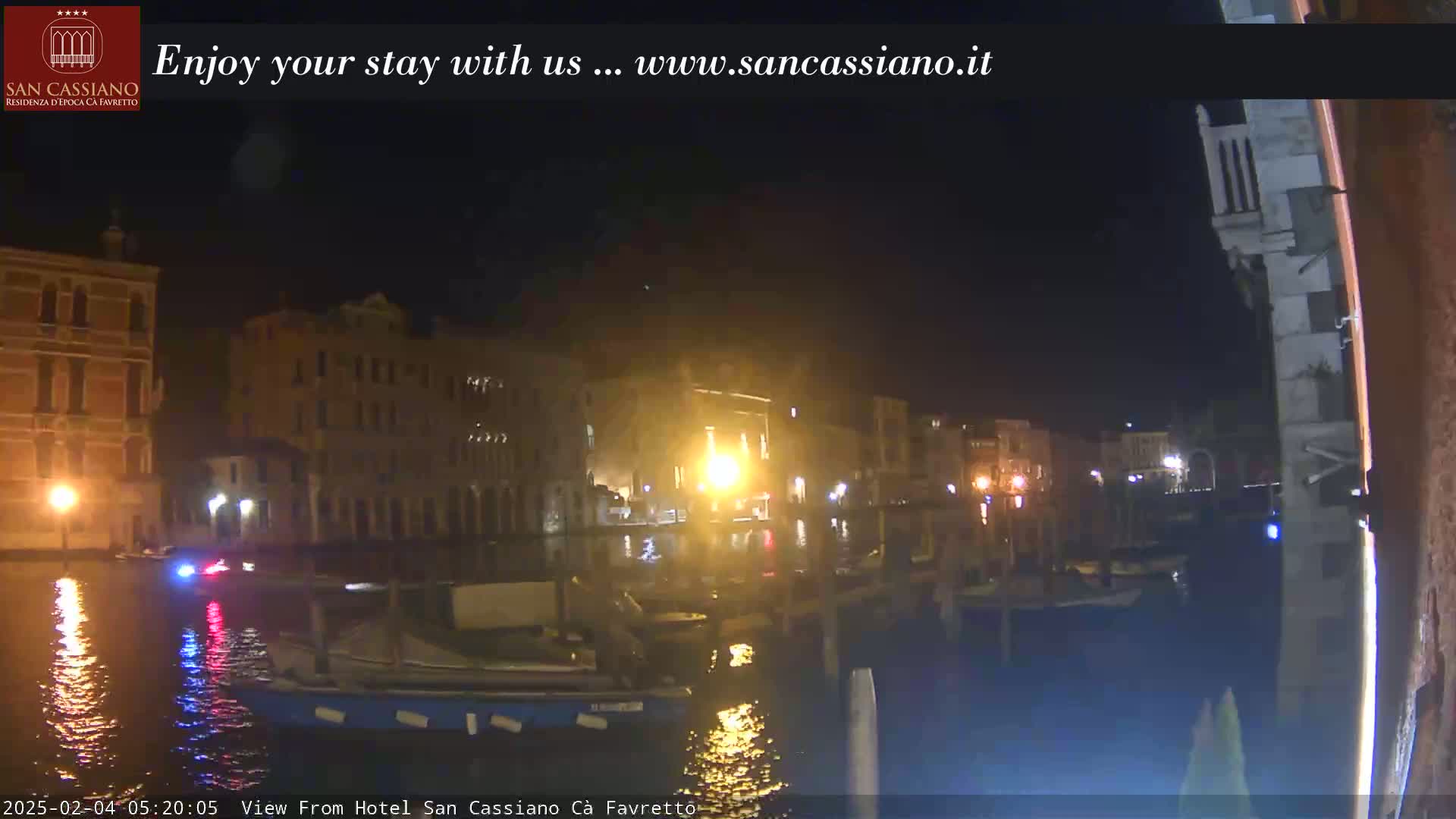 At night, several buildings line a canal, with boats moored along the waterway.