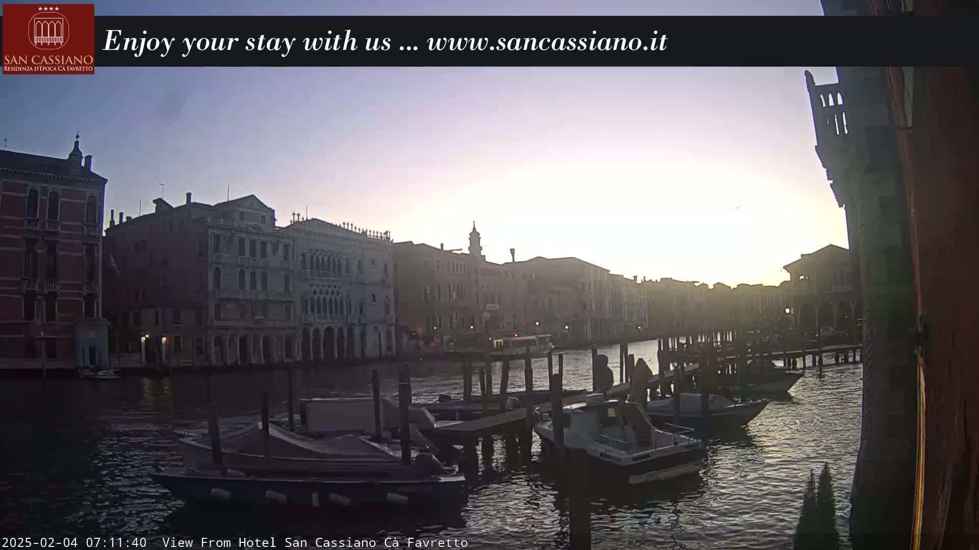 A sunrise illuminates a Venetian canal, where several boats are moored alongside historic buildings.