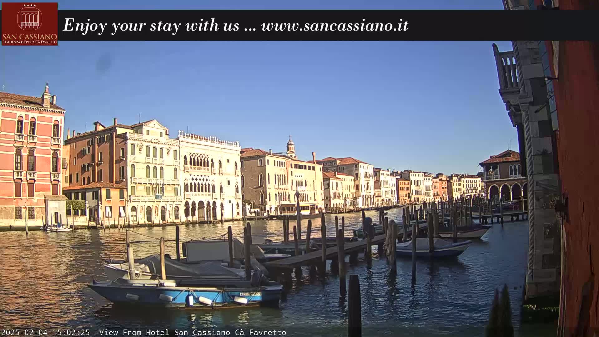 A sunny day in Venice shows a canal with several boats moored at wooden docks, and colorful buildings lining the waterway.