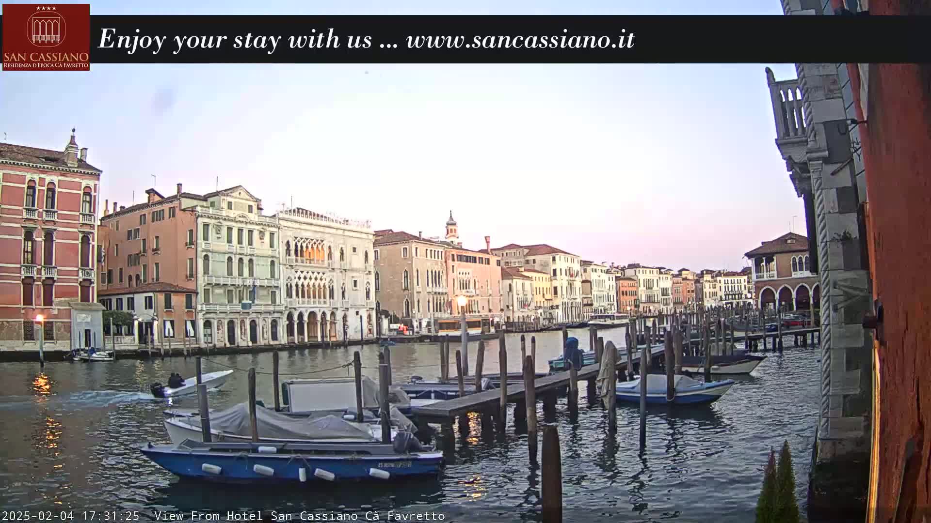 A tranquil canal scene in Venice shows several boats moored at a dock in front of colorful buildings under a pale, clear sky.