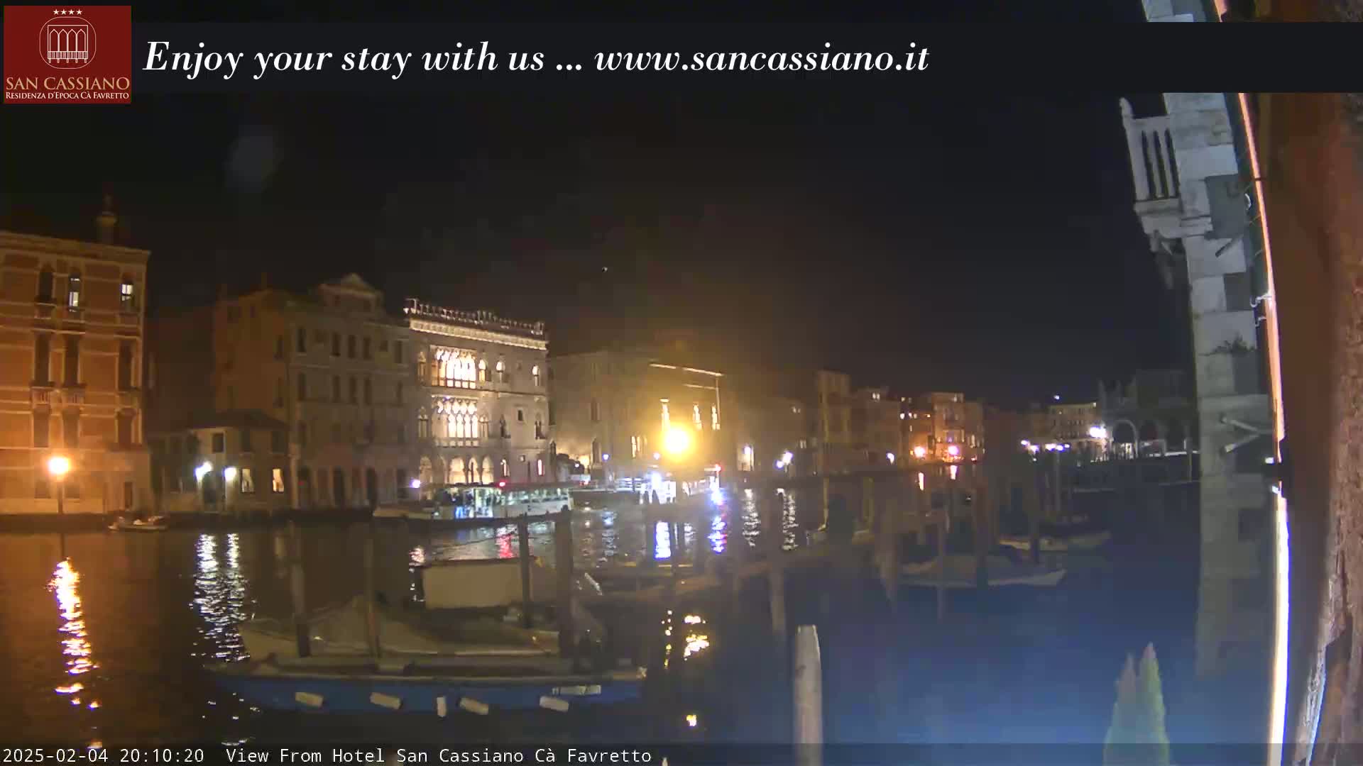 A nighttime view of a canal in Venice, Italy, shows several buildings illuminated along the waterway, with boats moored nearby.
