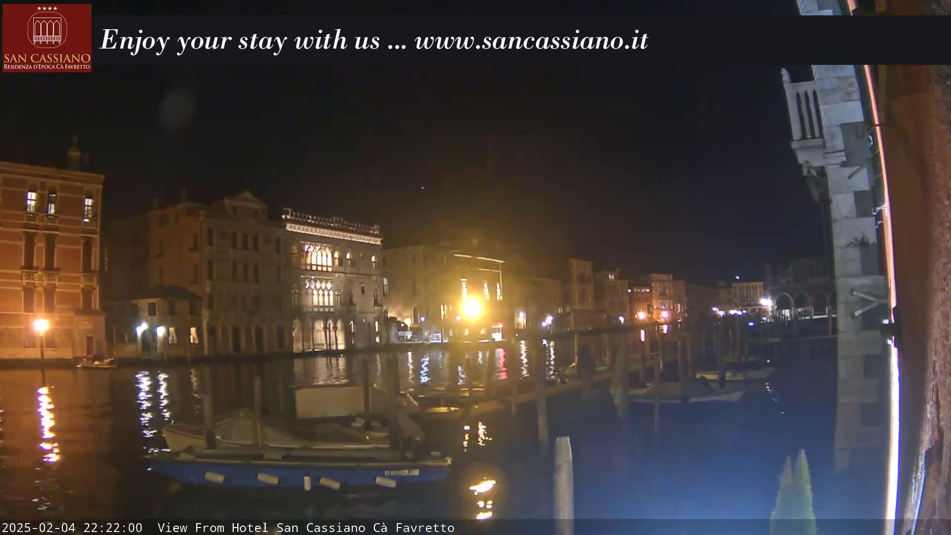 A nighttime view of a canal in Venice, Italy, showing several buildings illuminated along the waterway with boats moored at the edge.