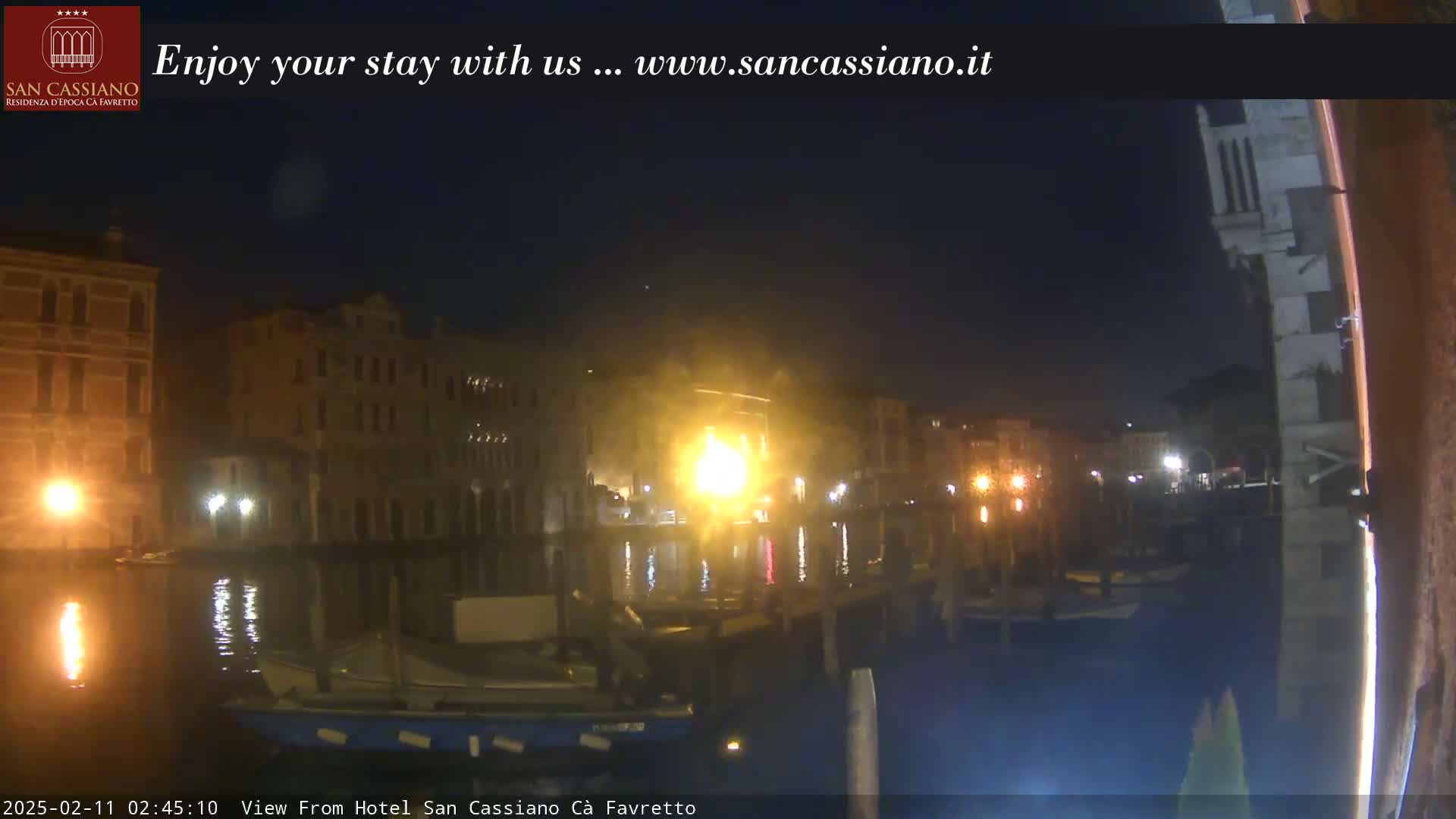 A nighttime view of a canal in Venice, Italy, shows buildings lit up along the water's edge with boats moored nearby.