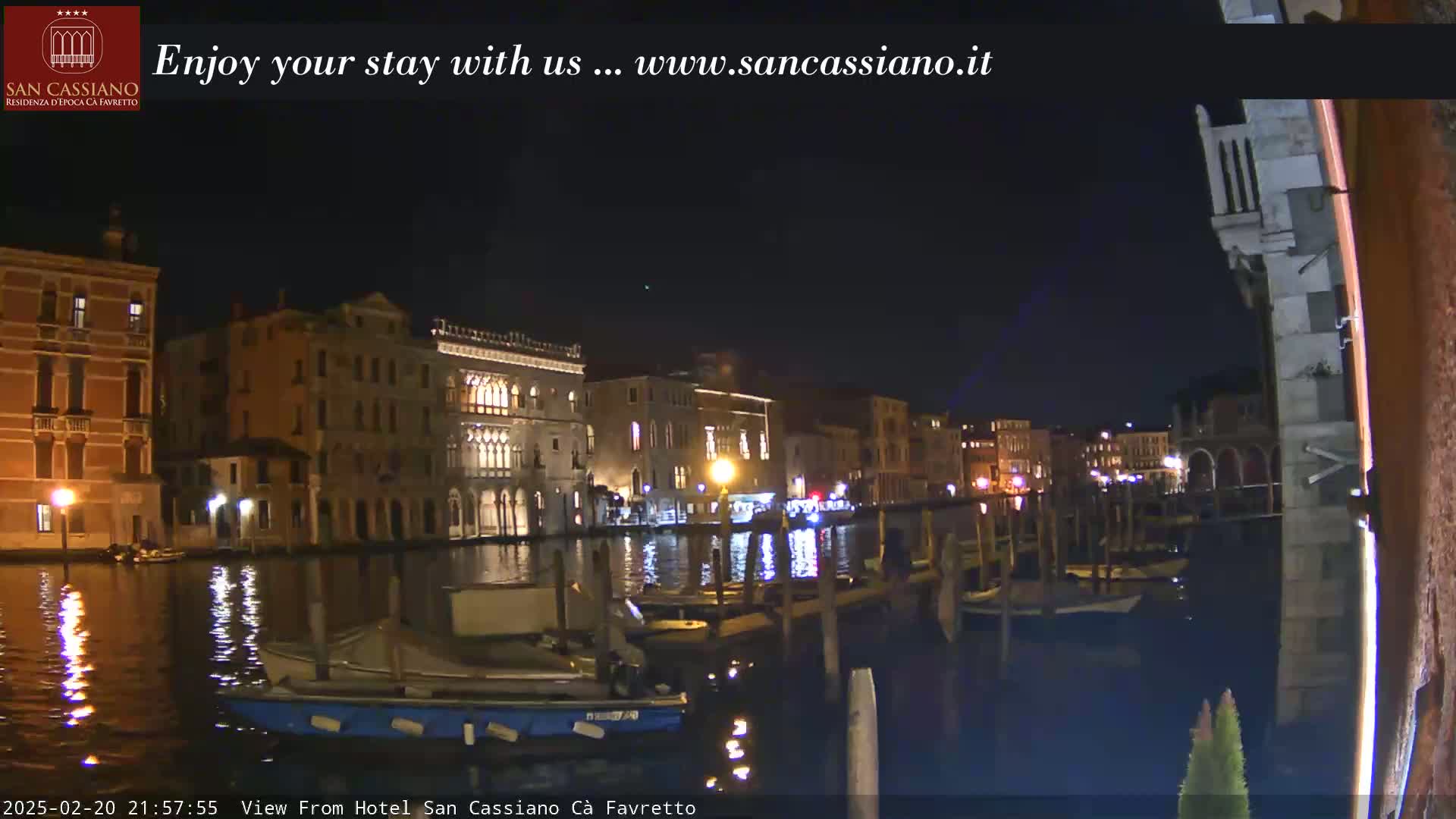 Nighttime view of a canal in Venice, Italy, with several boats moored along a stone embankment, illuminated buildings lining the waterway, and a calm, dark water surface reflecting the lights.