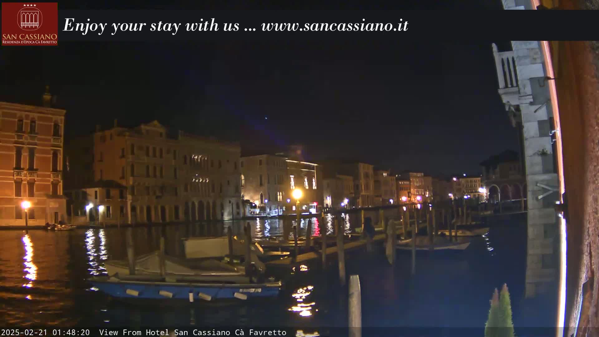 A nighttime view of a canal in Venice, showing several buildings lit up along the waterway and boats moored at the side.