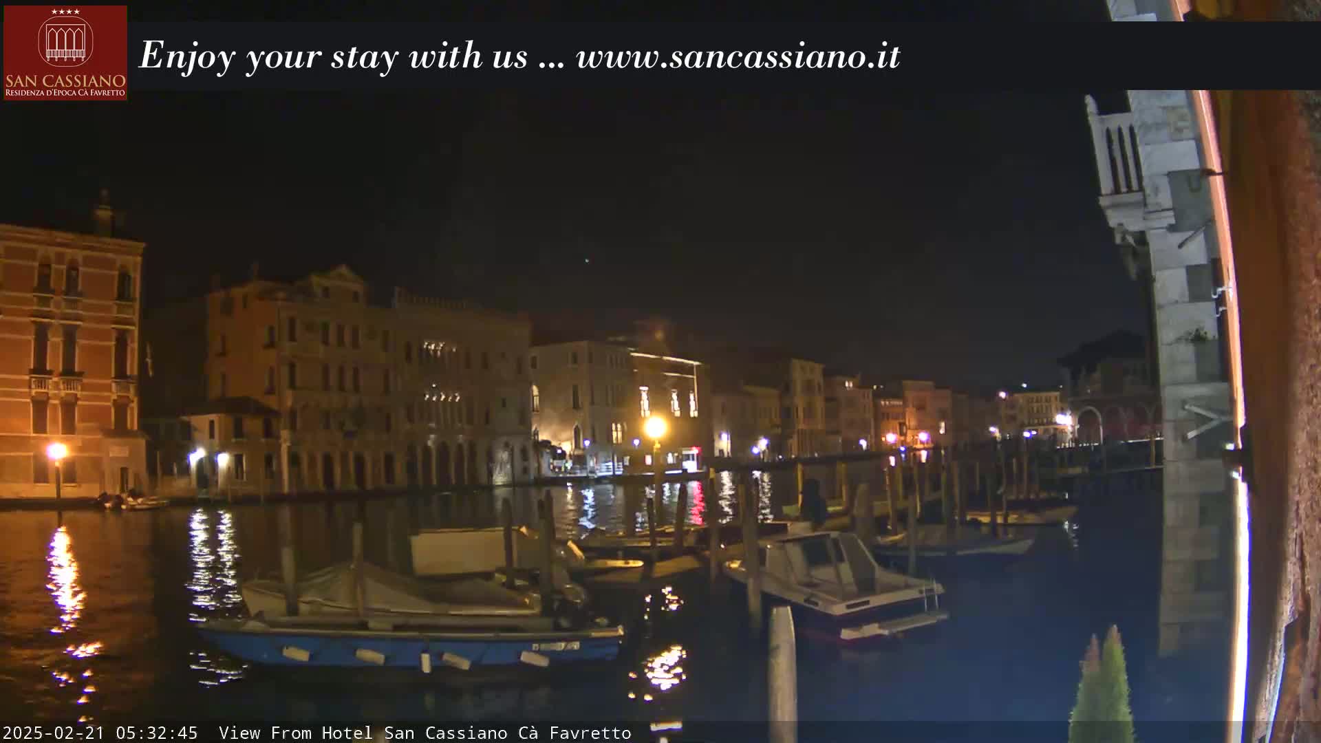 A nighttime view of a canal in Venice, Italy, shows several moored boats and illuminated buildings along the waterway.