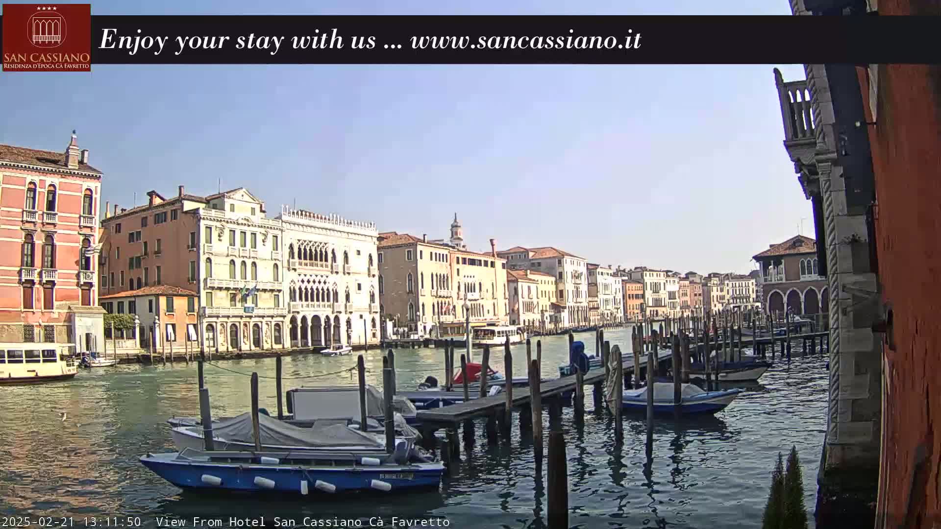 A sunny view of a canal in Venice, Italy, shows several moored boats and historic buildings lining the waterway.