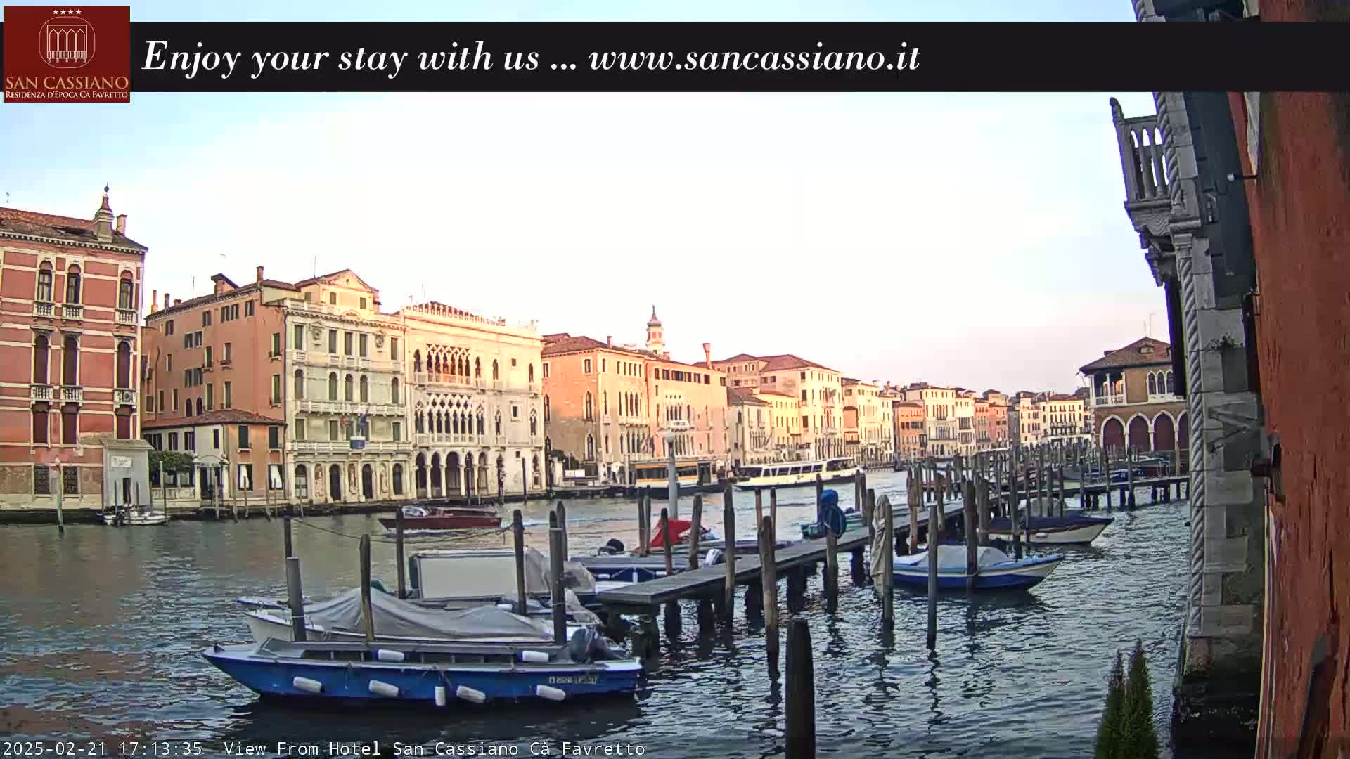 A calm canal in Venice shows several moored boats near a dock with multiple buildings lining the waterway under a clear, light-colored sky.