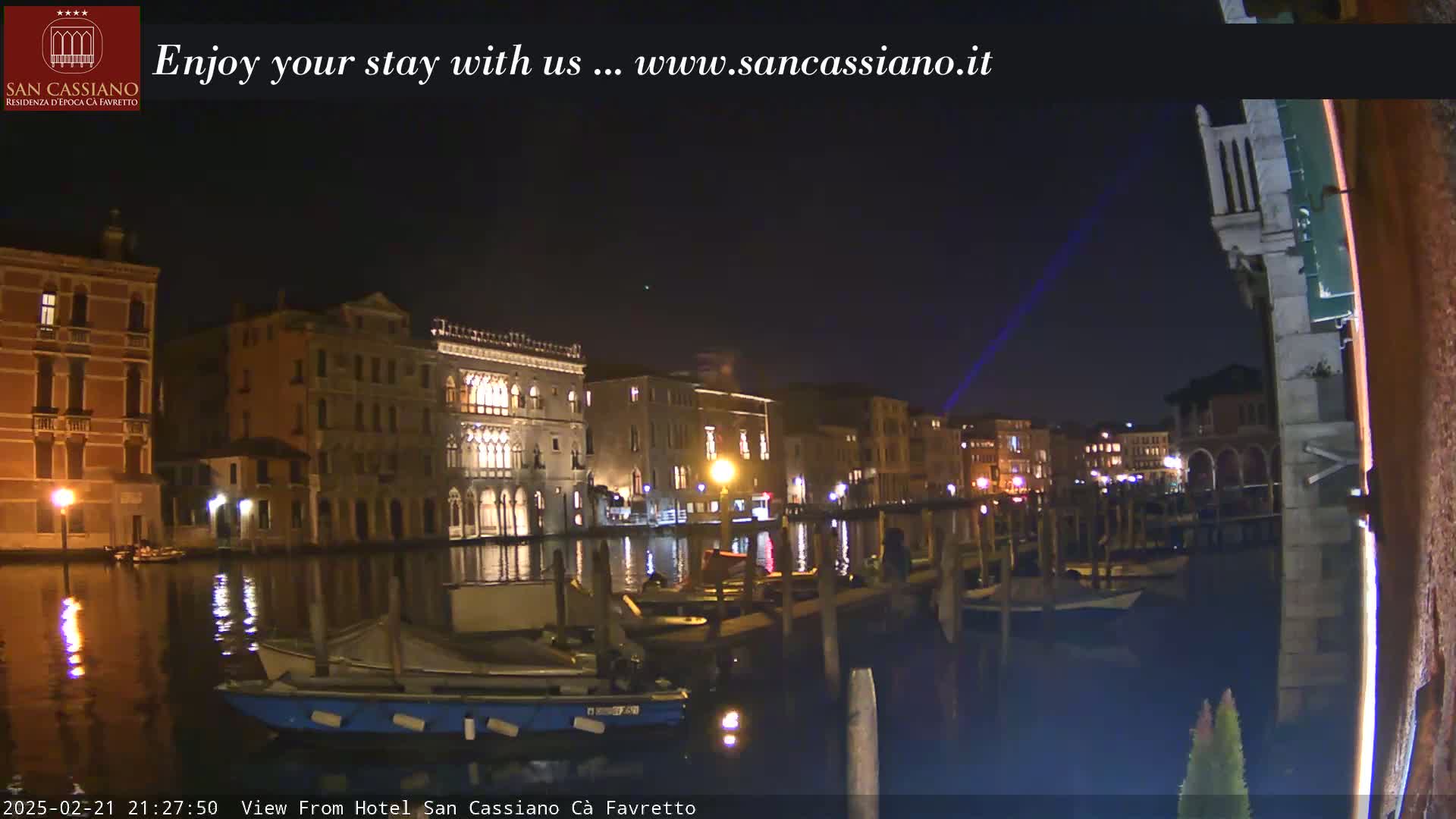 A nighttime view of a canal in Venice, showing illuminated buildings along the waterfront and several boats moored at the edge, under a clear dark sky with a laser beam visible.