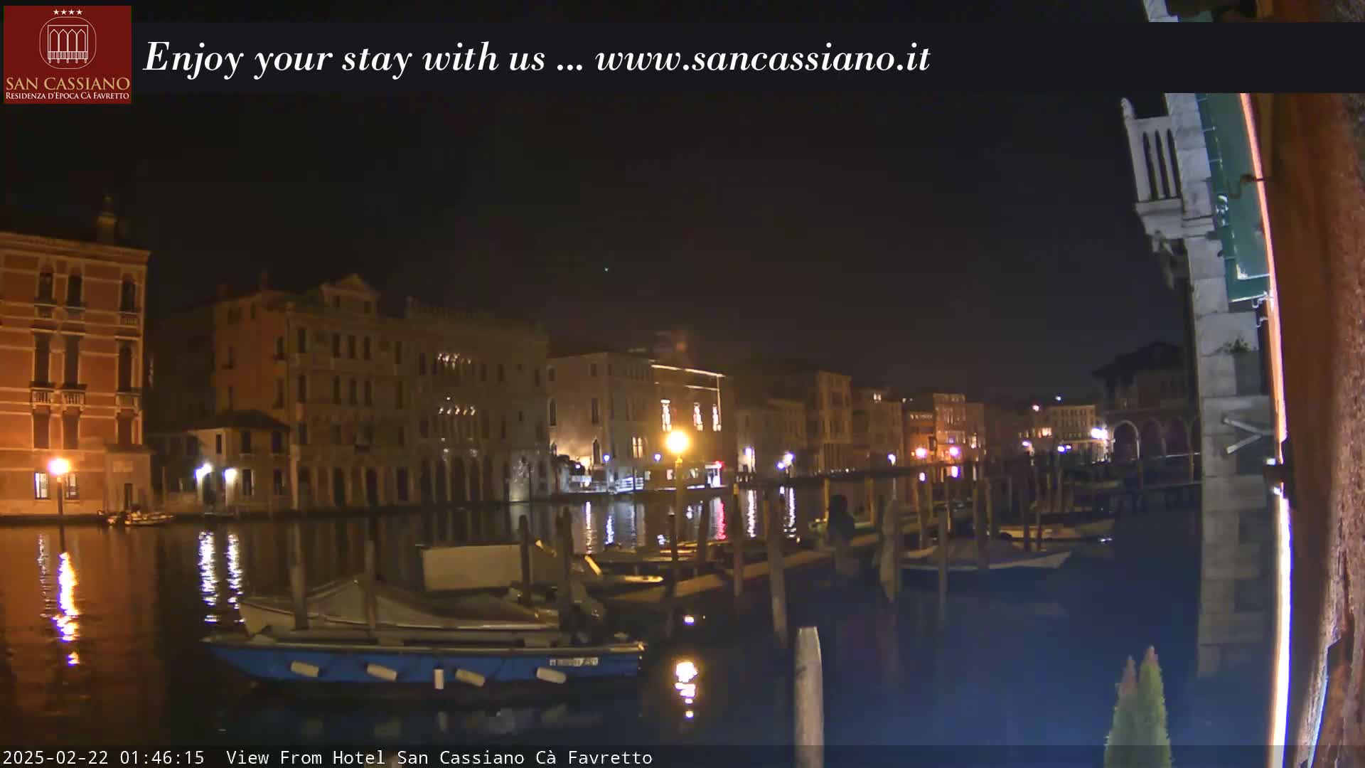 A nighttime view of a canal in Venice, Italy, shows several buildings lit up along the waterway with boats moored along the edge.