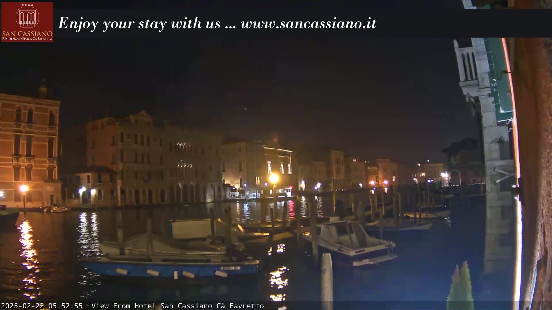 A nighttime view of a canal in Venice, Italy, showing several moored boats and illuminated buildings along the waterway under a clear, dark sky.