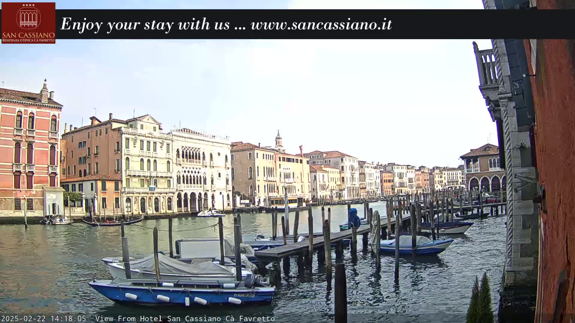 A canal in Venice, under a mostly clear sky, shows several docked boats and colorful buildings lining the waterway.