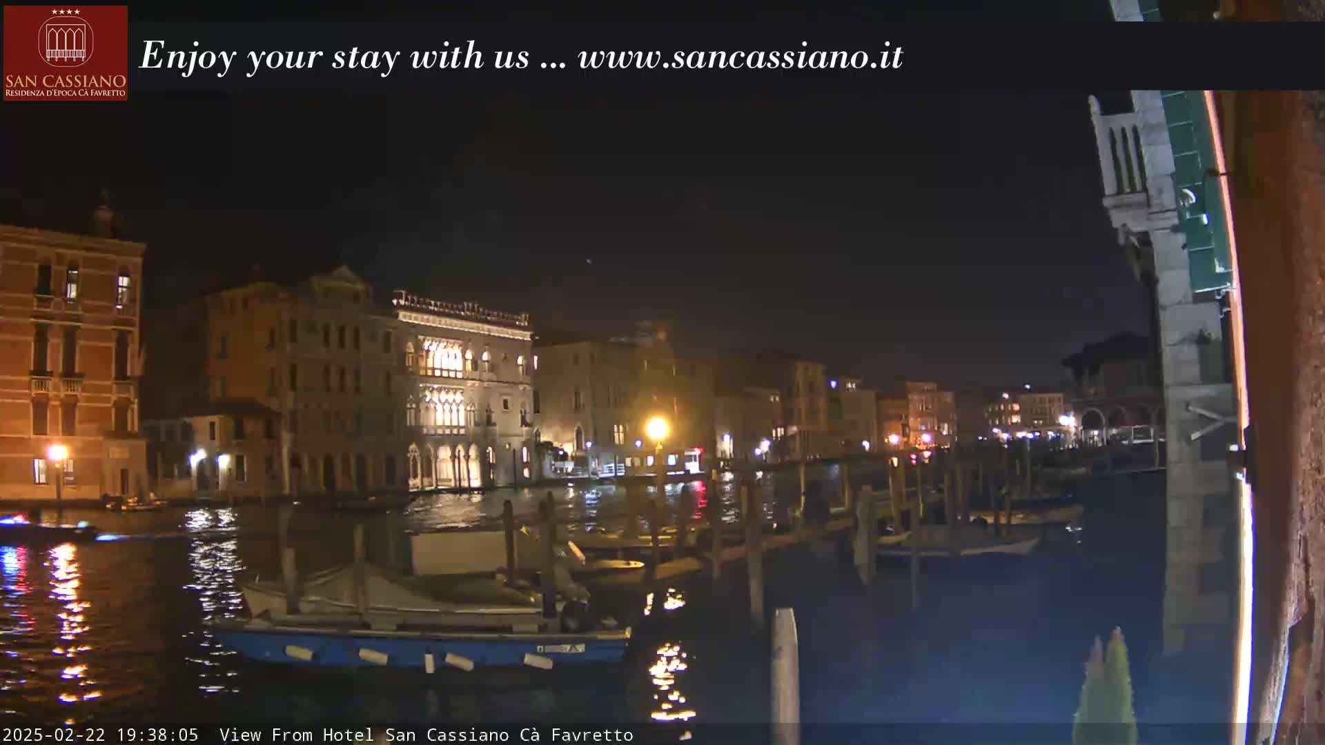 A nighttime view of a canal in Venice, Italy, shows several buildings lit up along the waterway, with boats moored along the docks.