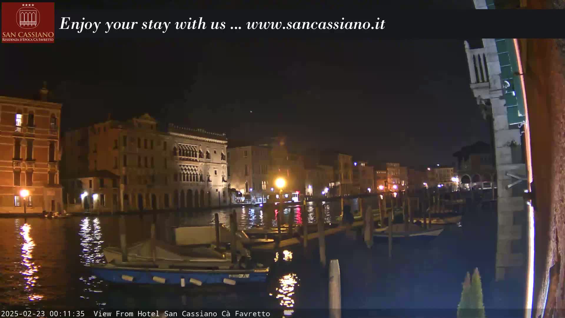 A nighttime view of a canal in Venice, with several boats moored along a dock, and buildings illuminated along the waterway.