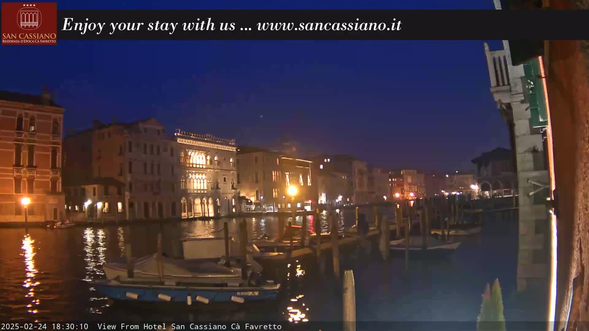 A nighttime view of a canal in Venice, Italy, shows several buildings lit along the waterway with boats moored at the edge.
