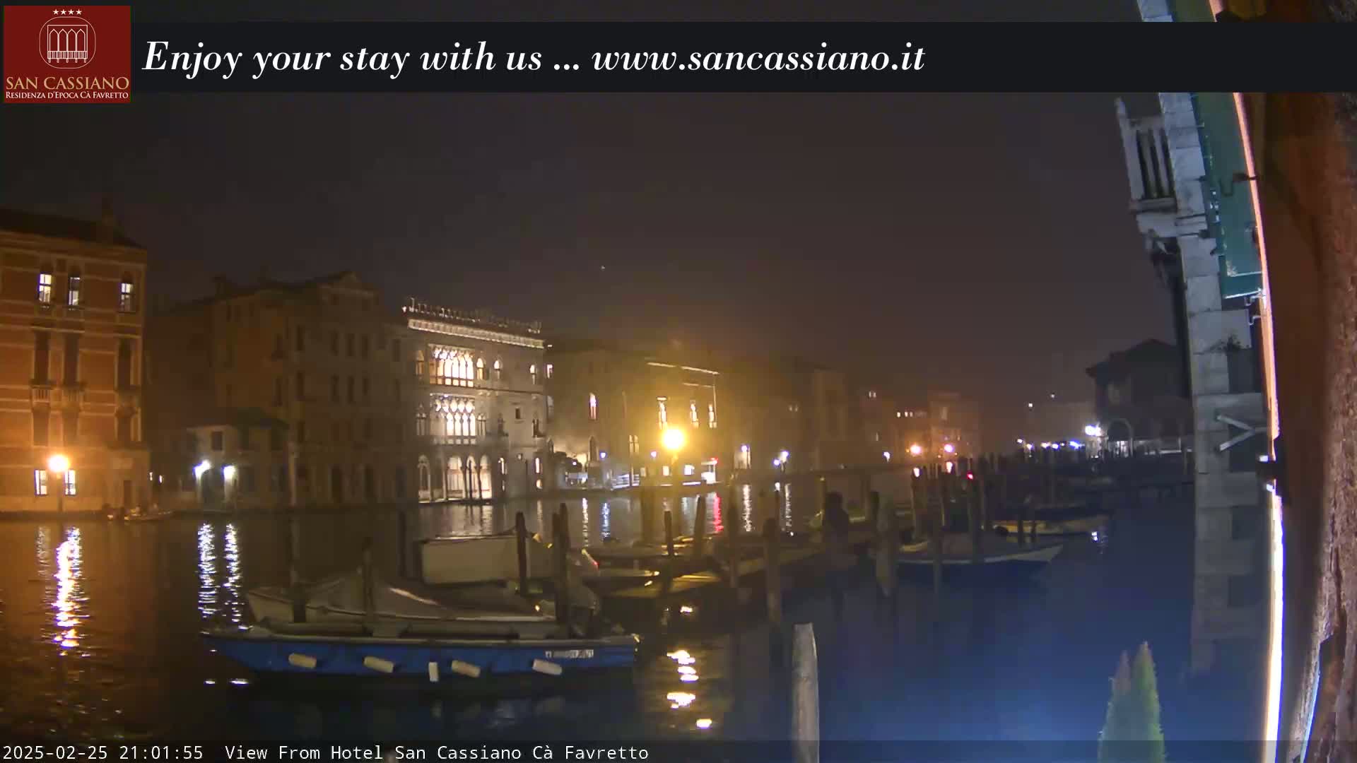 A nighttime view of a canal in Venice, showing illuminated buildings and moored boats under a dark, slightly hazy sky.