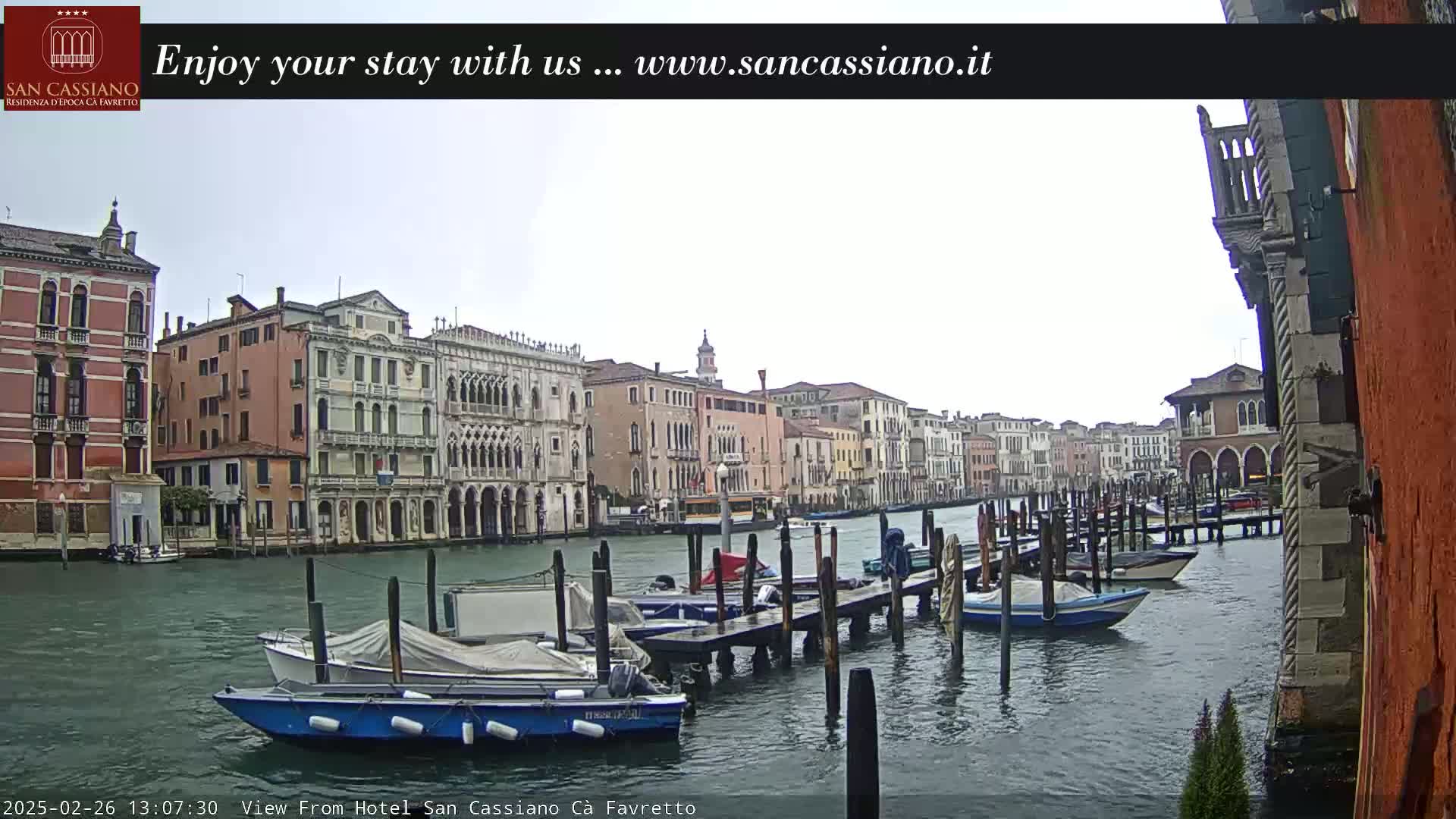 Overcast skies loom over a Venetian canal, where several boats are moored at a dock in front of historic buildings.