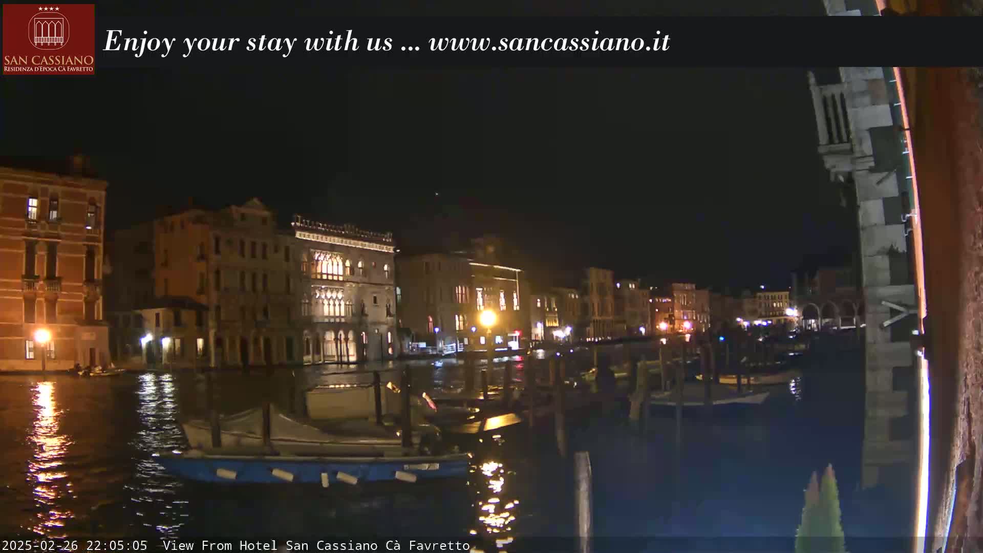 A nighttime view of a canal in Venice, Italy, showing several buildings illuminated along the waterway, with boats moored on the water.