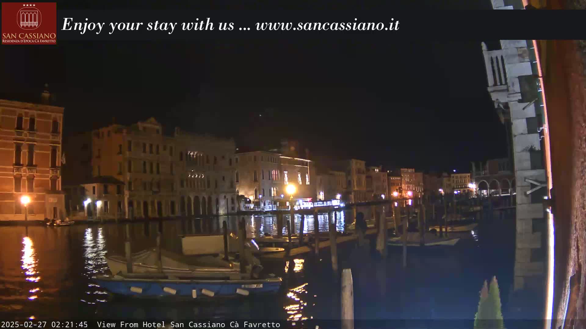 A nighttime view of a canal in Venice, with several buildings lit along the waterway and boats moored along the banks.