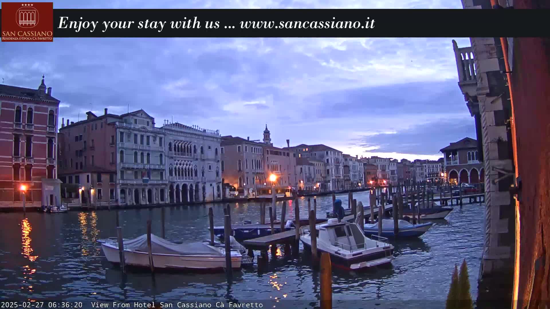 A twilight view of a Venetian canal, showing several moored motorboats and buildings lining the waterway under a partly cloudy sky.