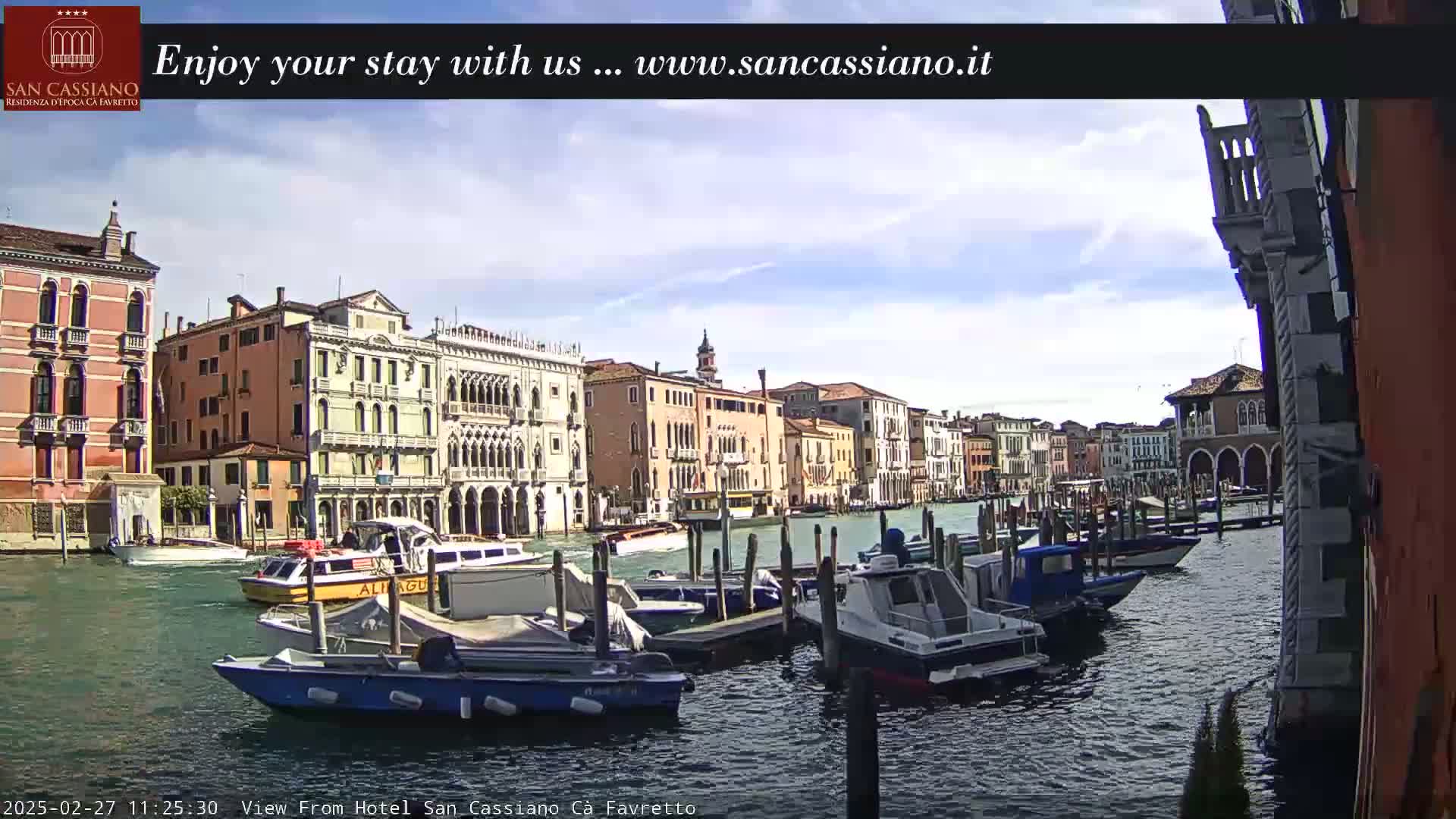 A canal in Venice, Italy, with several moored boats and buildings lining the waterway under a partly sunny sky.