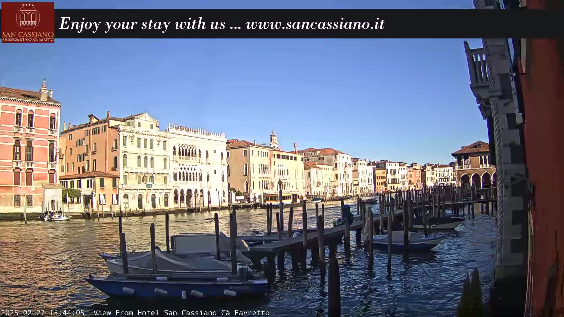 Several colorful buildings line a canal in Venice, under a clear blue sky, with several boats moored at a dock.