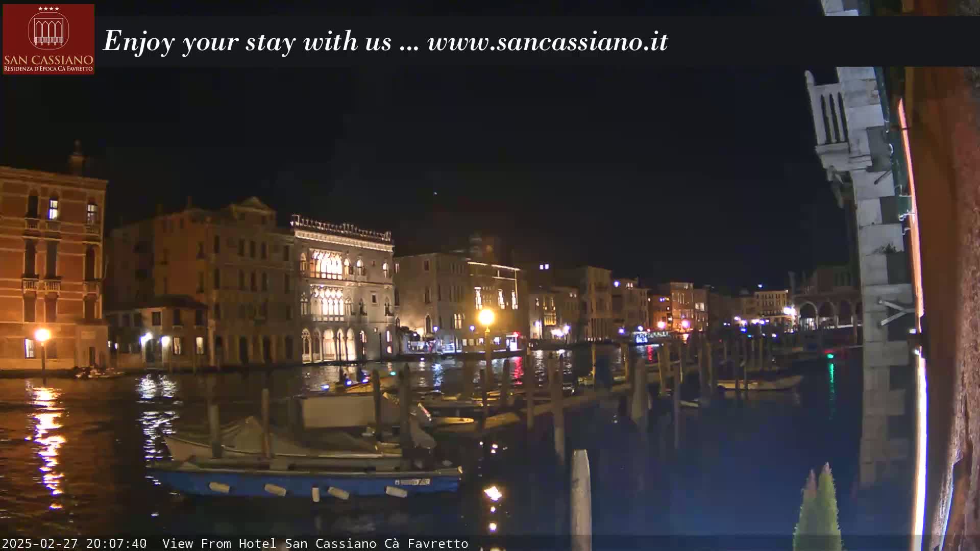 A nighttime view of a canal in Venice, Italy, showing several buildings illuminated along the waterway, with several small boats moored along the canal.