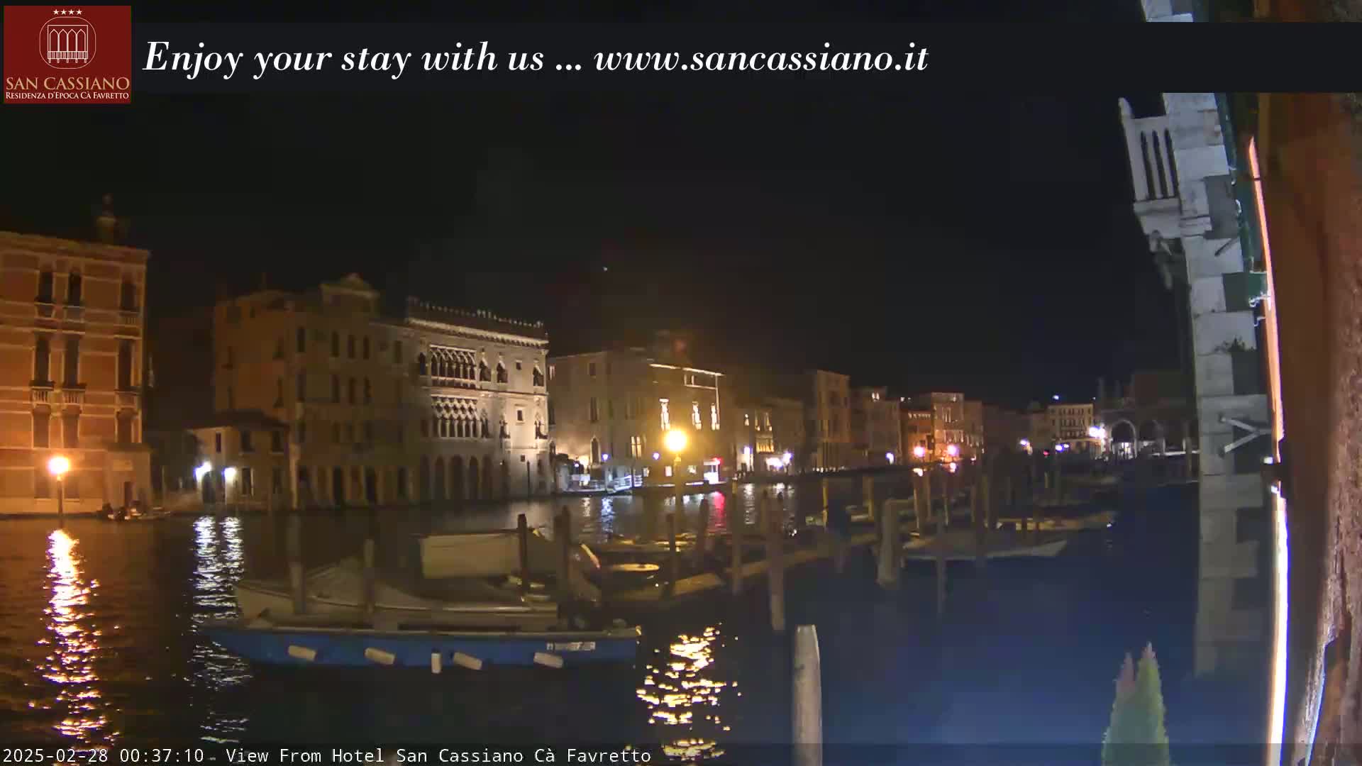 A nighttime view of a canal in Venice, with several buildings lit up along the waterway and small boats moored at the edge.