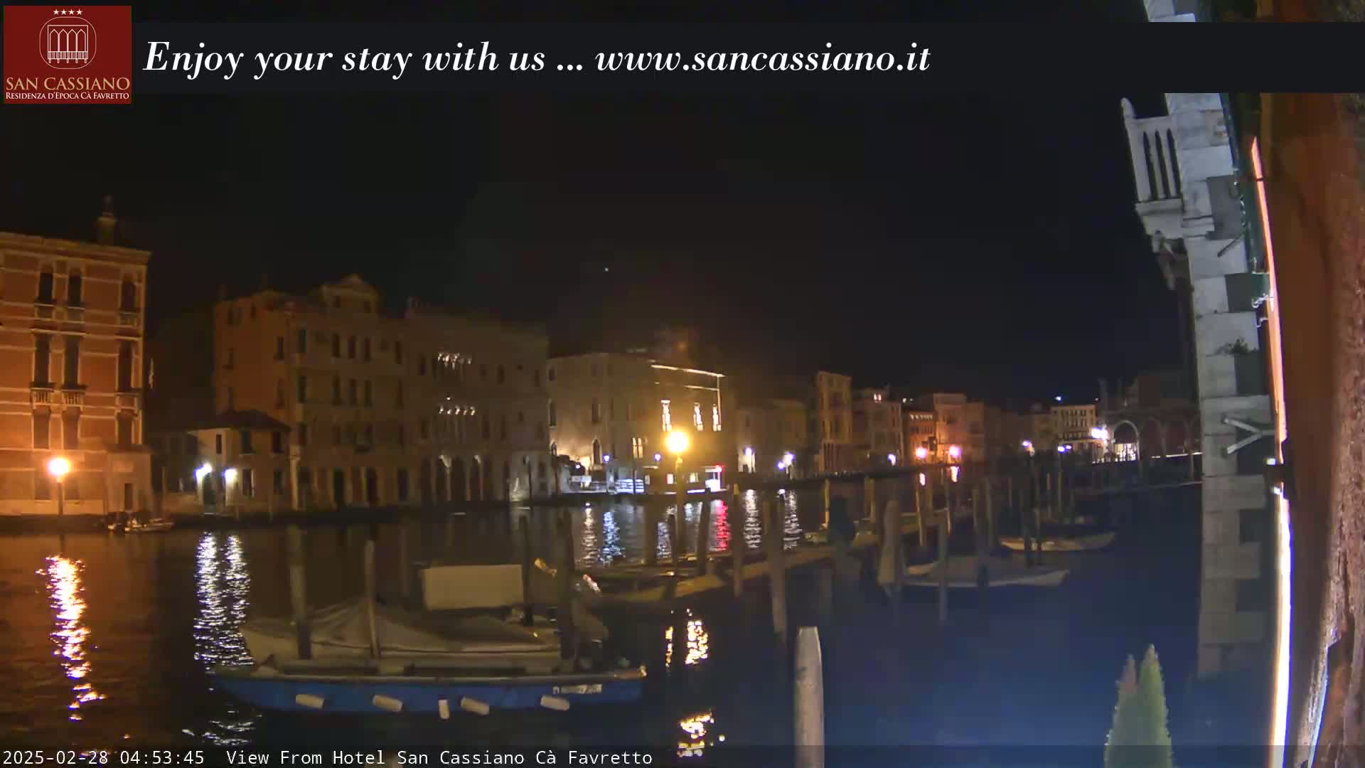 A nighttime view of a canal in Venice, Italy, with illuminated buildings lining the waterway and several boats moored along the banks.