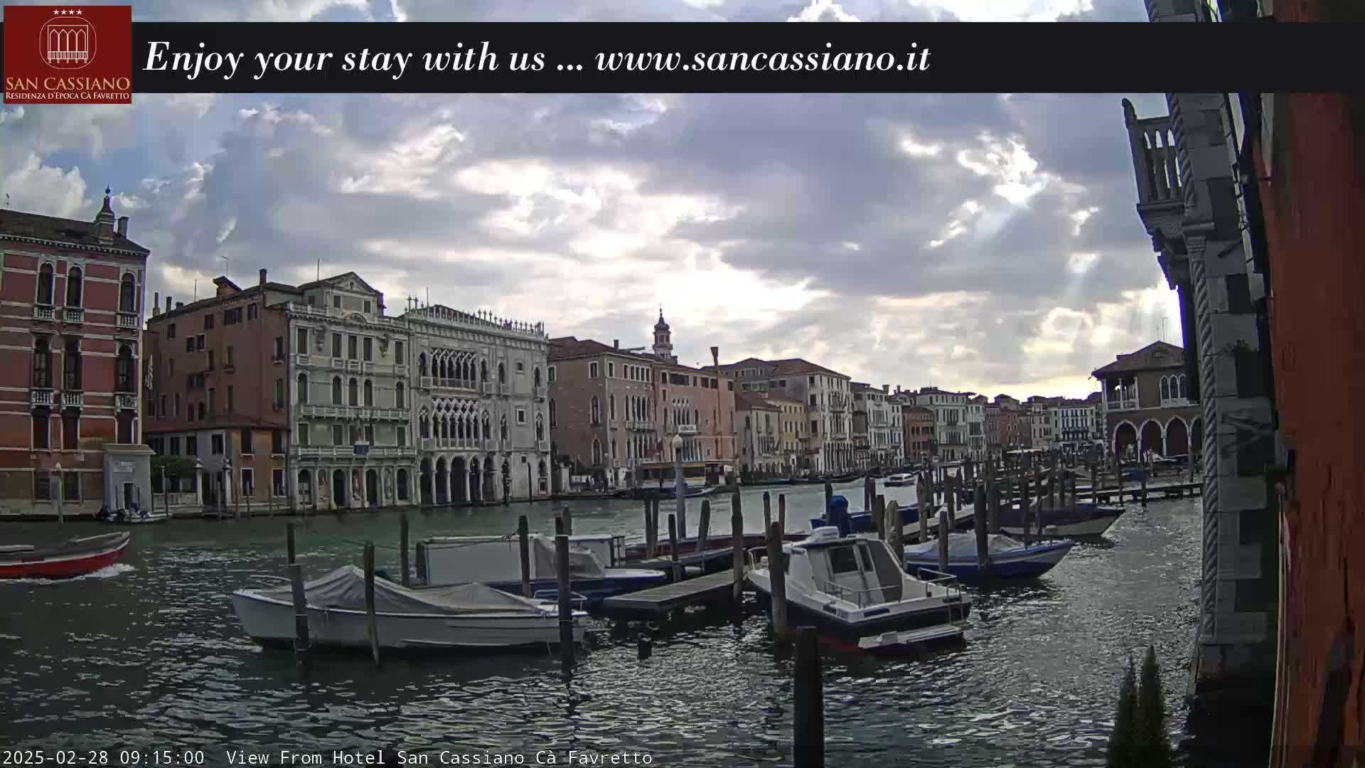 A canal in Venice, under a mostly cloudy sky, is lined with moored boats and historic buildings.