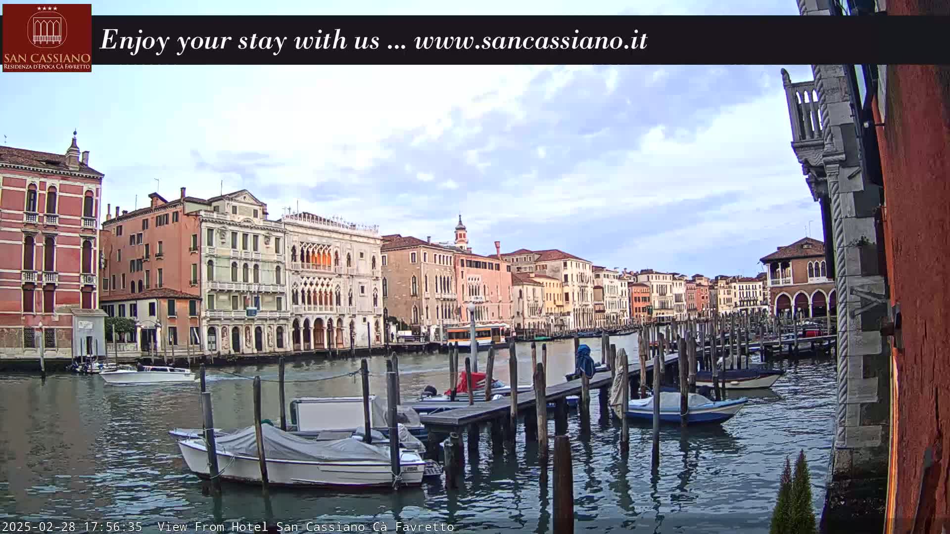 A canal in Venice, Italy, with several moored boats and colorful buildings lining the waterway under a partly cloudy sky.