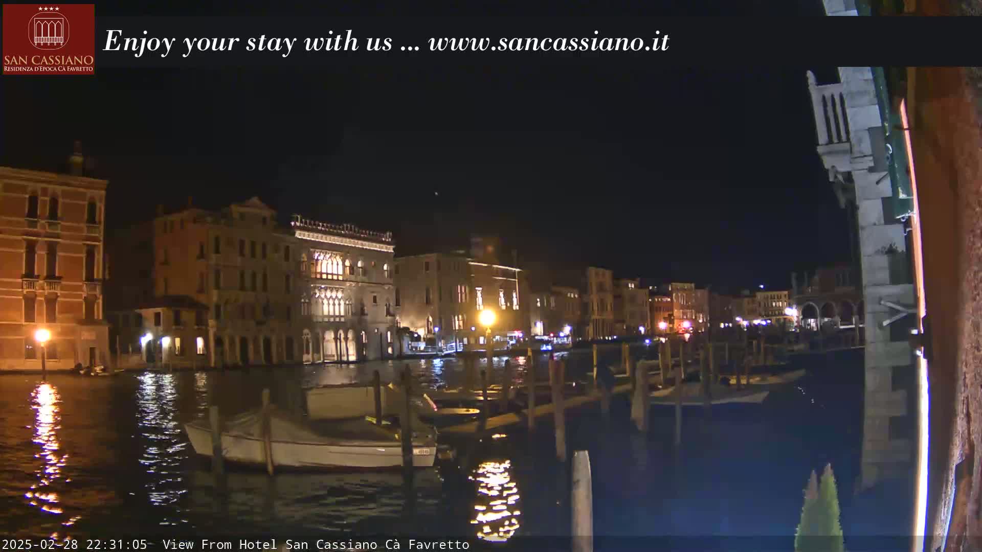 A nighttime view of a canal in Venice, Italy, shows several buildings illuminated along the waterfront with boats moored at the edge.