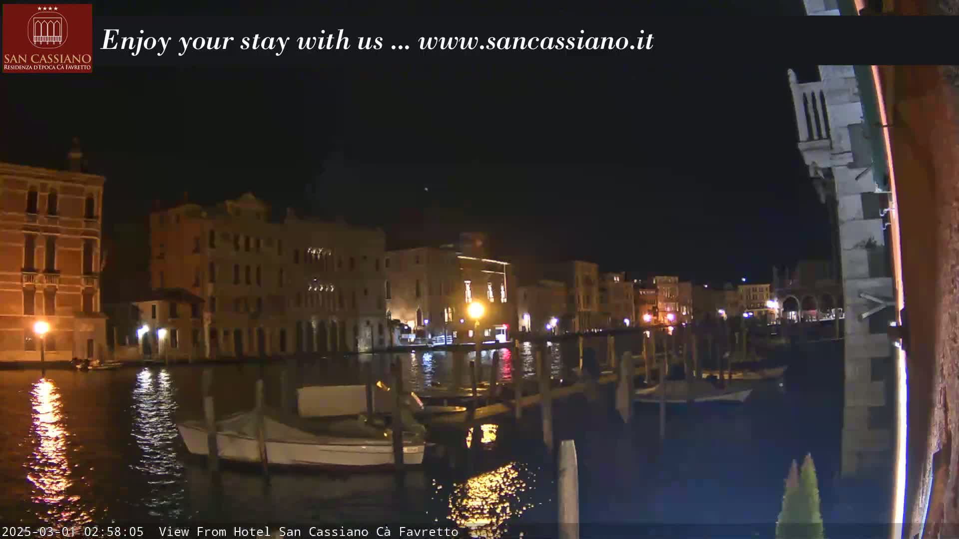 At night, several illuminated buildings line a calm canal, where small boats are moored.
