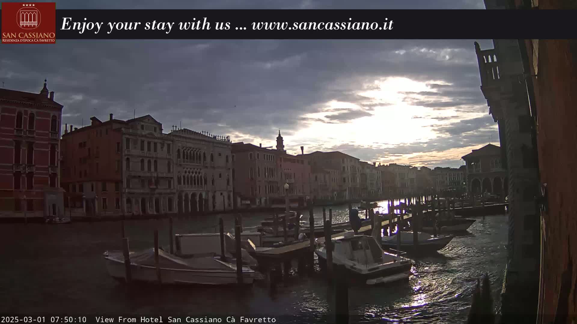 Several moored boats are visible on a canal lined with historic buildings at sunrise under a partly cloudy sky.