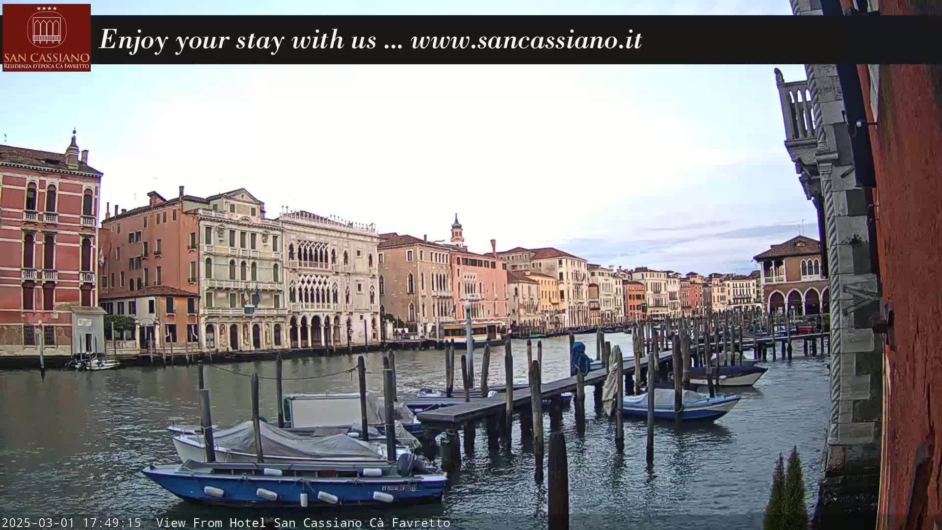 A canal in Venice, Italy, shows several moored boats and buildings along the waterway under a partly cloudy sky.