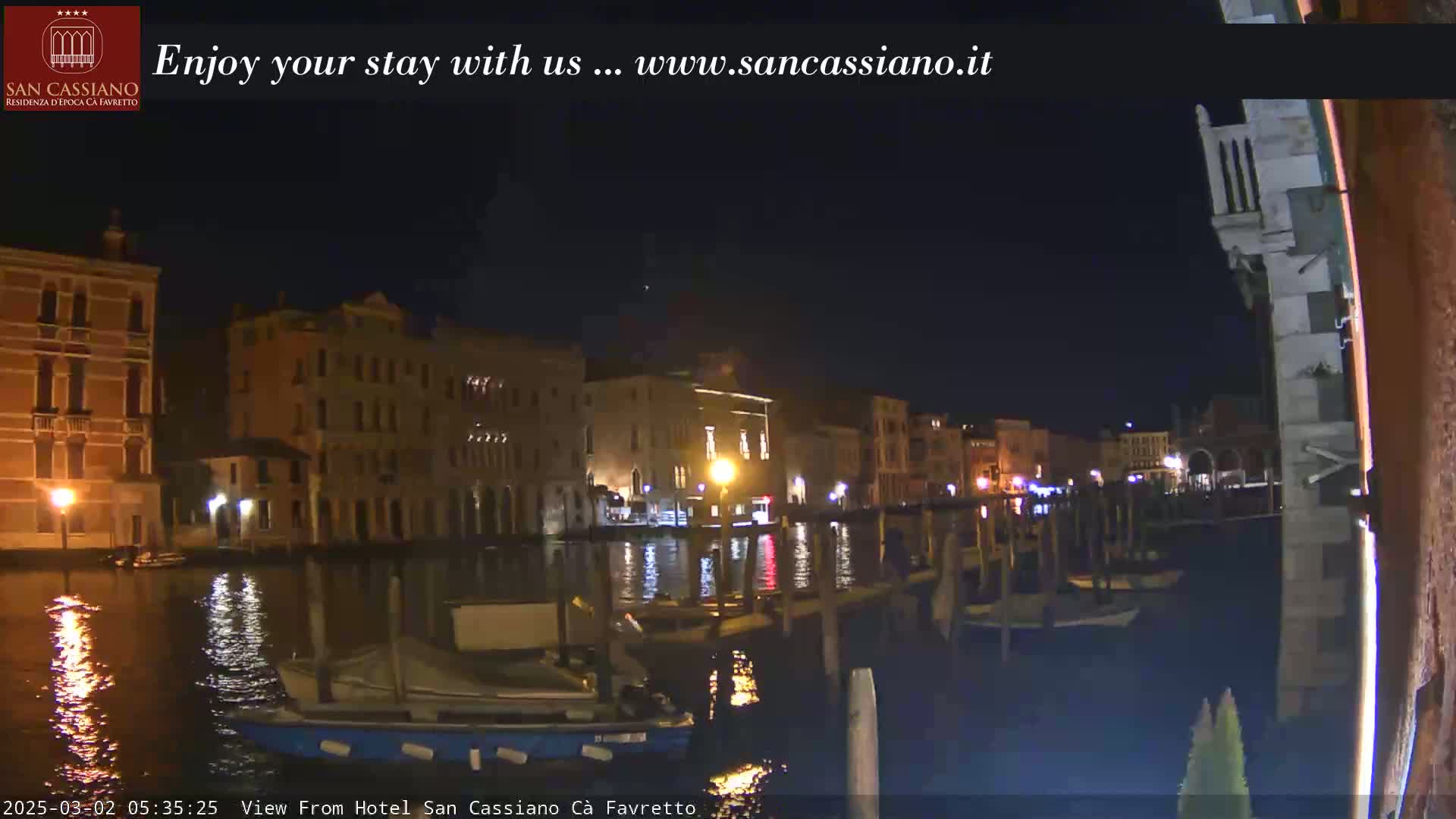 Nighttime view of a canal in Venice, Italy, with illuminated buildings lining the waterway and boats moored along the edges.